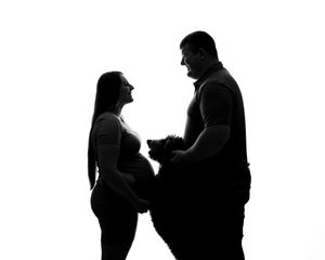A silhouette of an expecting couple holding their small dog against a white background.