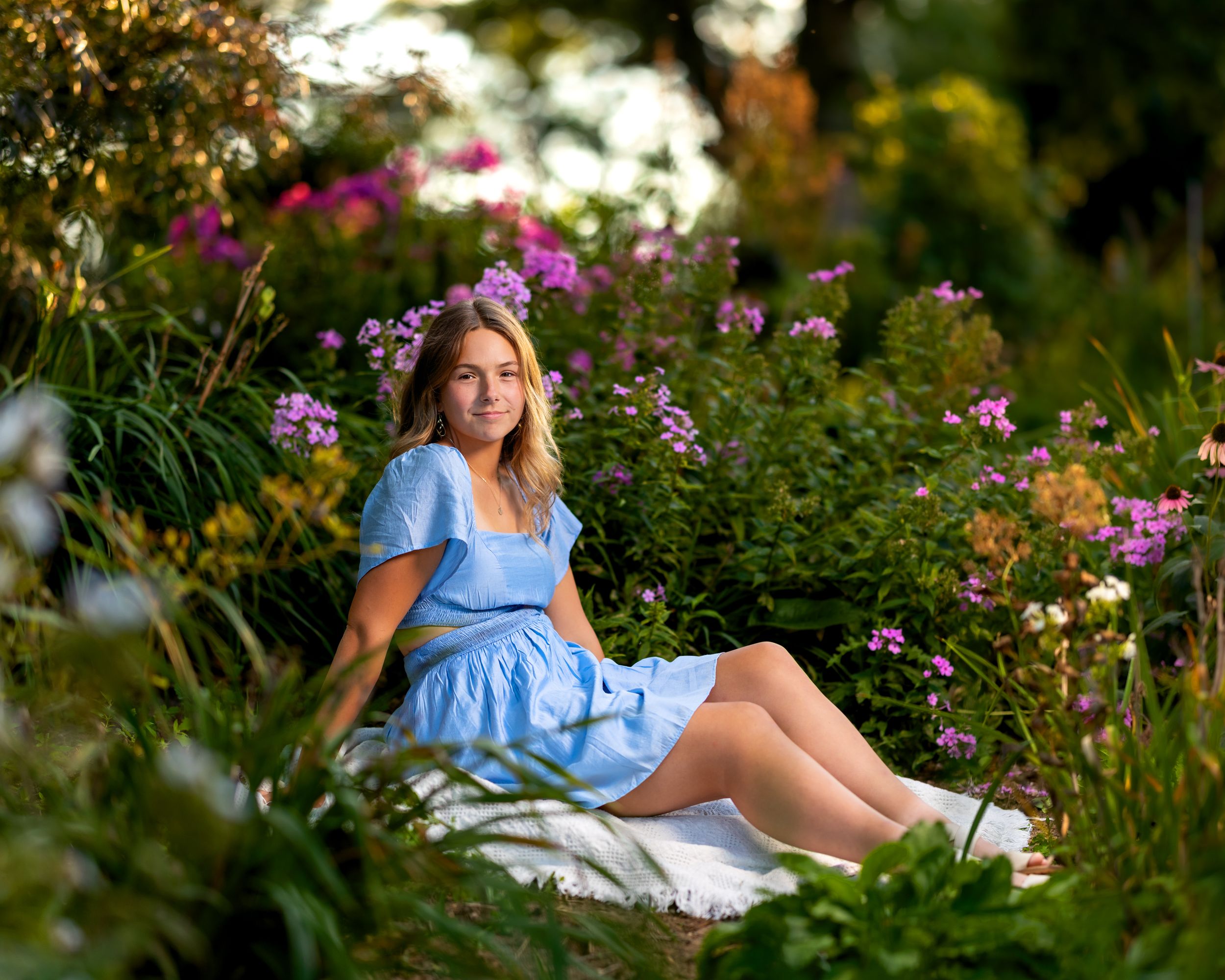 Young woman in blue dress sitting among colorful garden flowers during golden hour photography session.