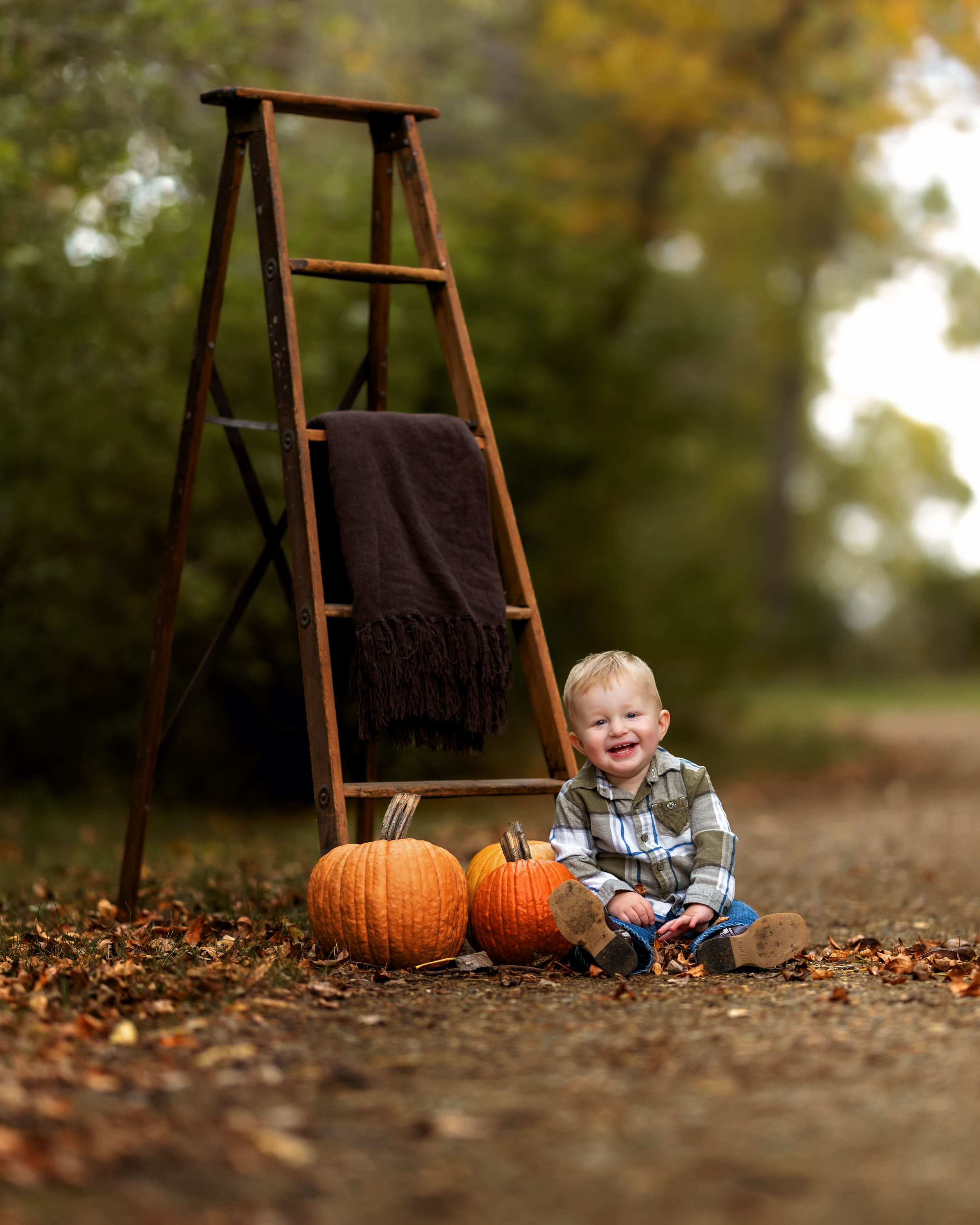 Smiling toddler sitting outdoors beside pumpkins and a rustic wooden ladder with a dark blanket in an autumn setting.