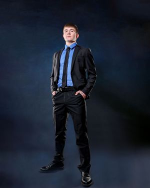 Young man in a black suit with blue shirt and tie posing confidently against a dark studio background.