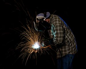 Welder in plaid shirt and protective mask creating bright sparks while welding metal in a dark workshop.