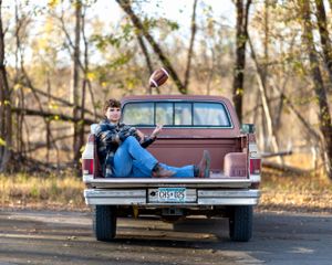 Teen boy relaxing in the bed of a vintage red pickup truck tossing a football in an autumn outdoor setting.