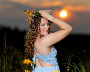 Young woman with curly hair wearing a wildflower crown poses at sunset in a field of sunflowers.