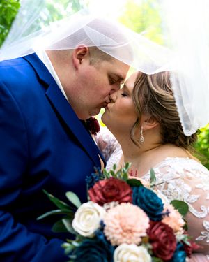 Bride and groom sharing a romantic kiss under a wedding veil with a colorful bridal bouquet in the foreground.