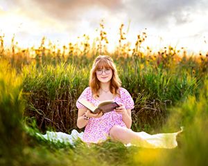 A young woman with glasses sits in a golden field at sunset, reading a book in a pink floral dress.