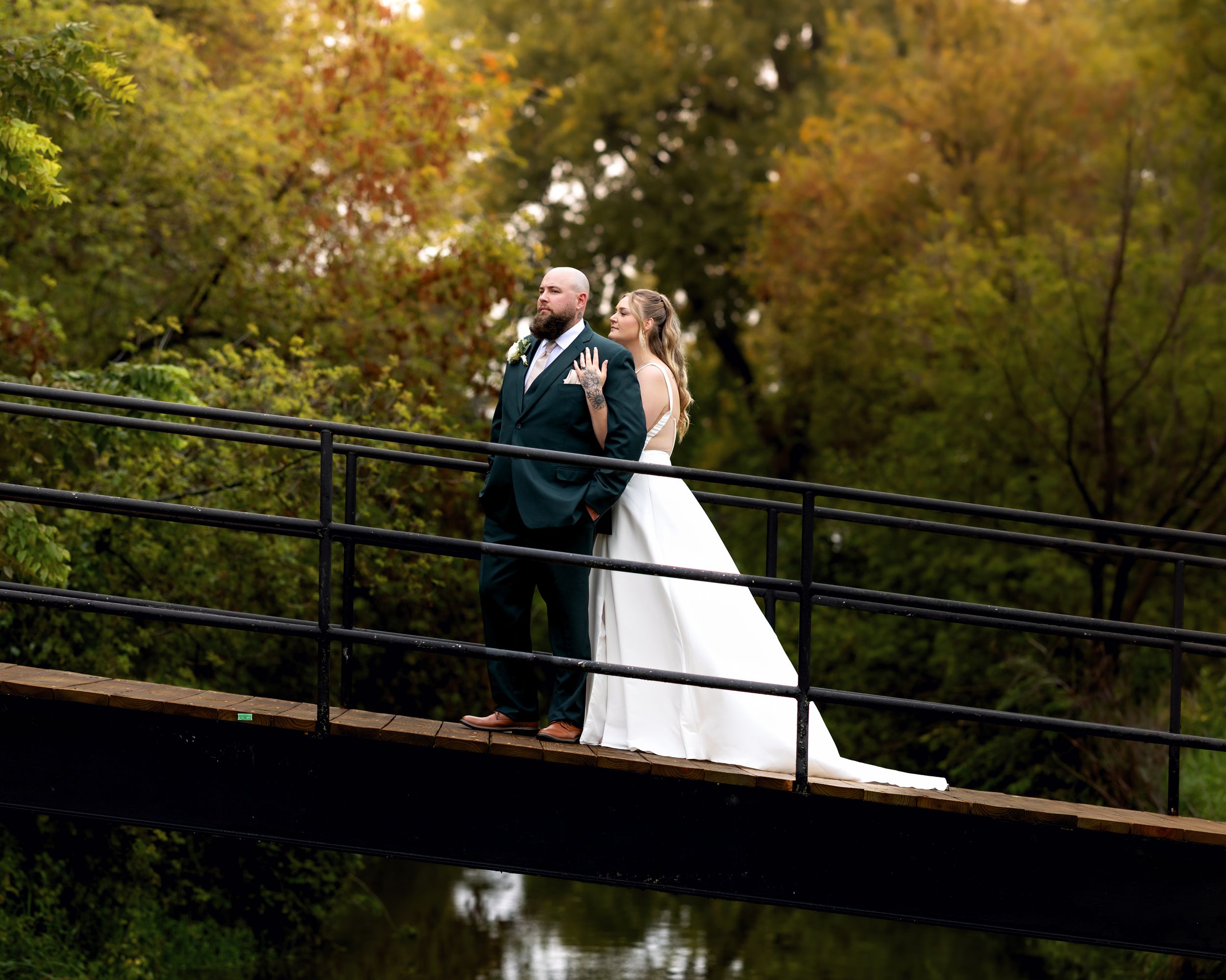 Bride and groom share a romantic moment on a bridge surrounded by lush autumn foliage near a serene river.