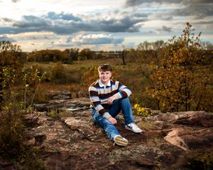 Teenage boy in striped polo sits on rocky terrain surrounded by autumn foliage under dramatic cloudy sky.