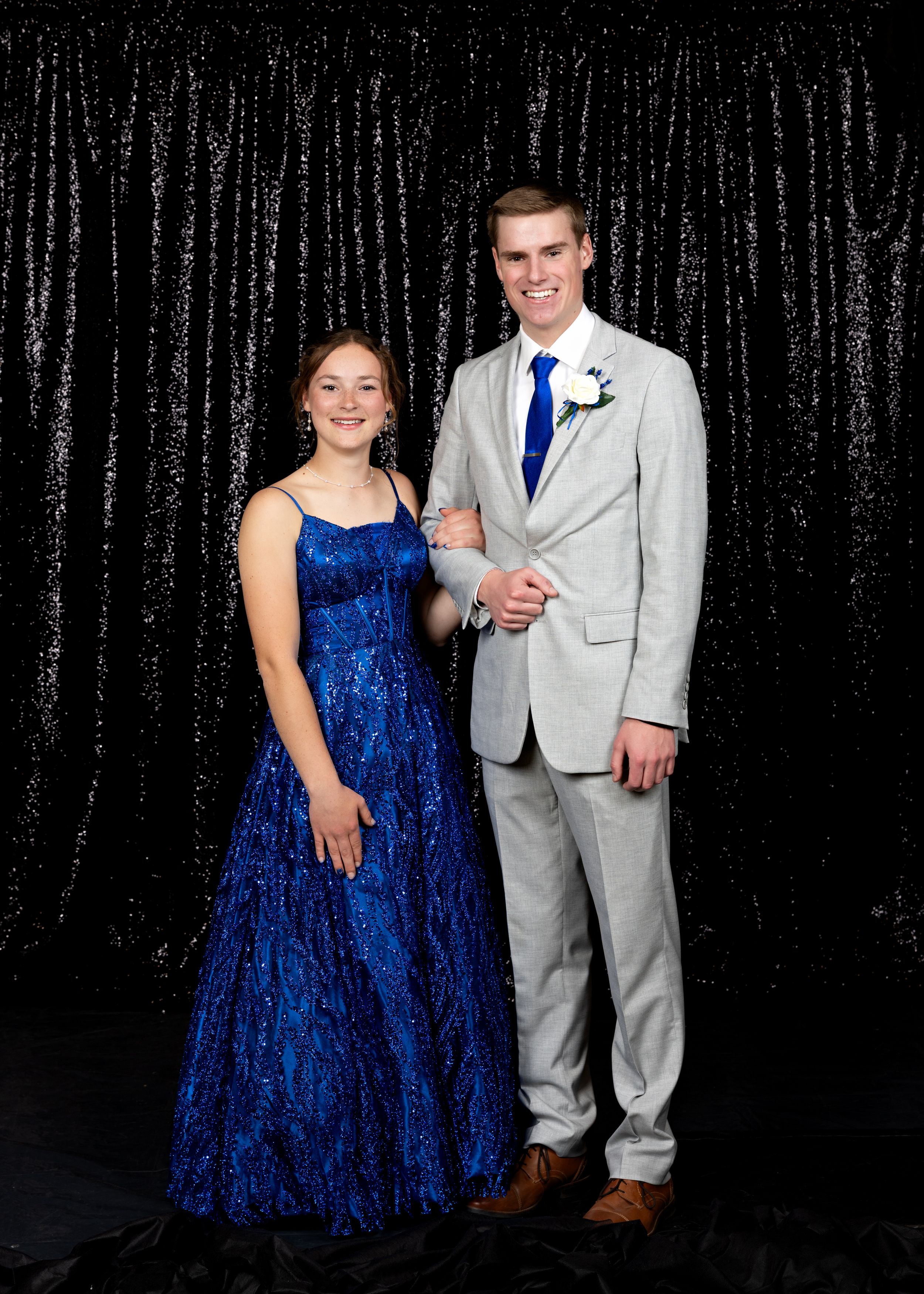 Young couple dressed in formal prom attire pose together in front of black sequin backdrop.