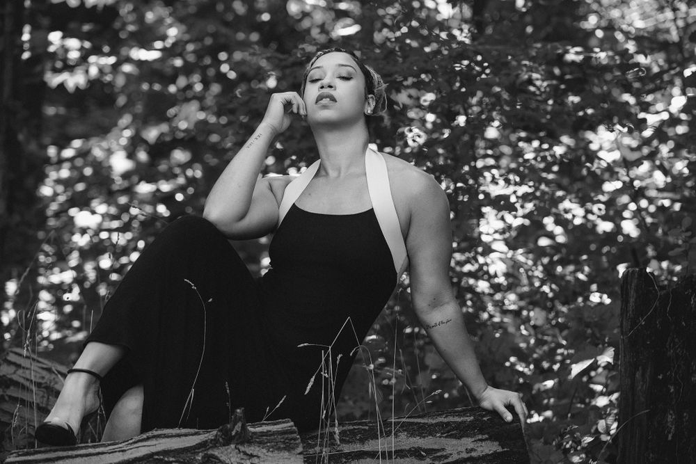 A black and white artistic portrait of someone in a black tank top looking up against a blurred nature backdrop.