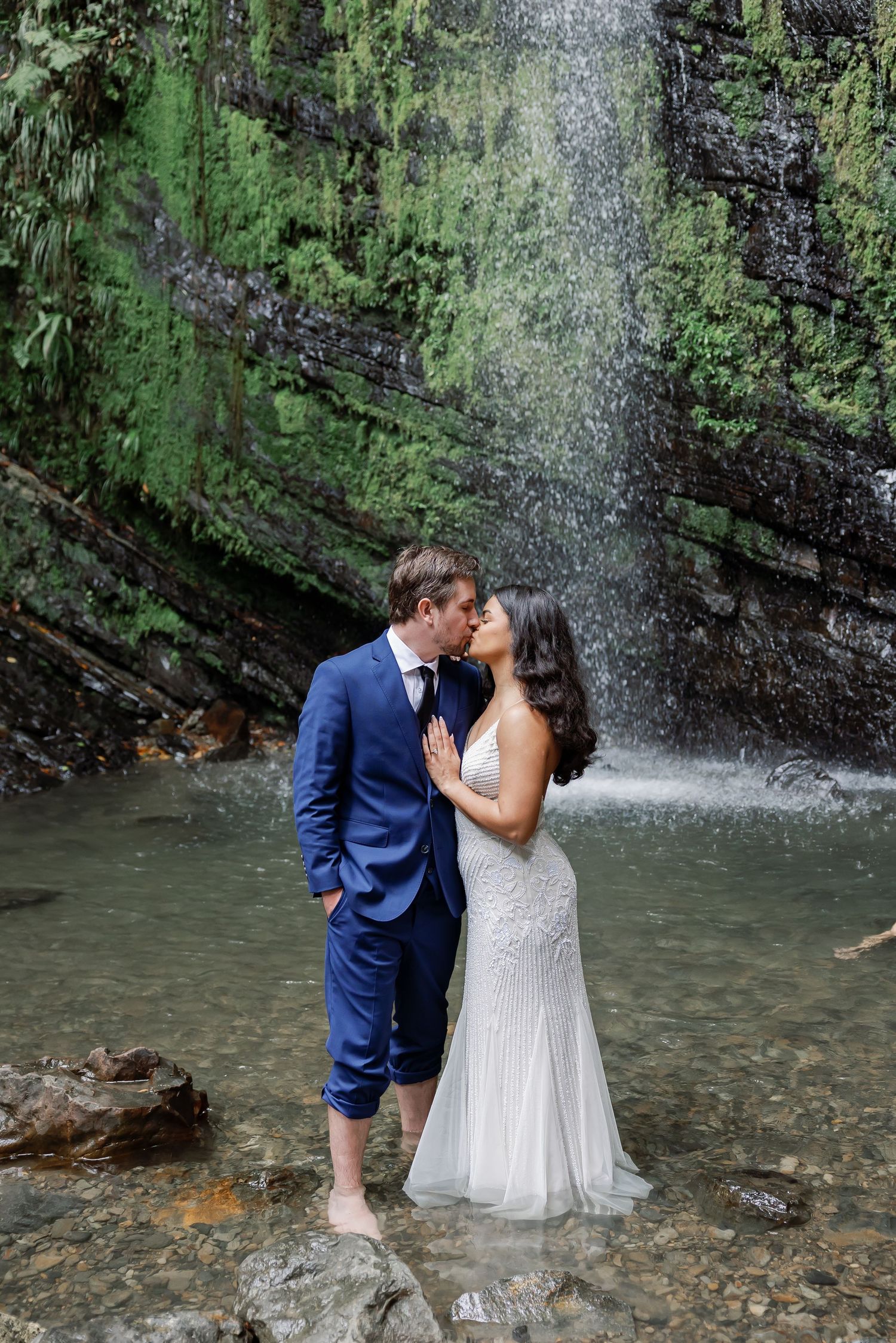 A bride and groom embrace in a tropical forest setting, with a waterfall in the background.