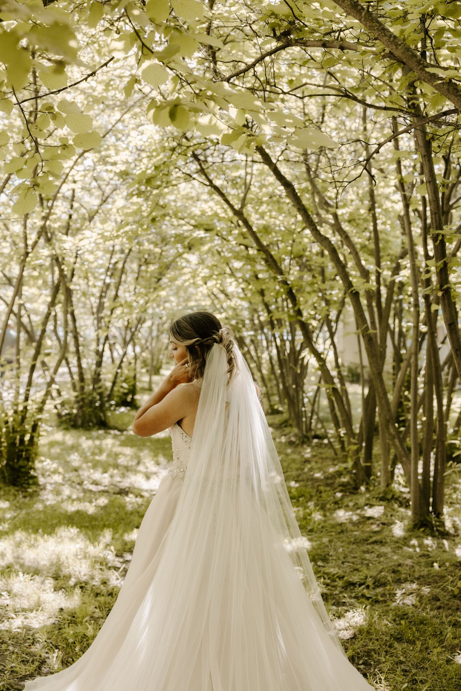 A bride in a flowing white wedding dress stands gracefully in a sunlit forest setting with dappled light through trees.