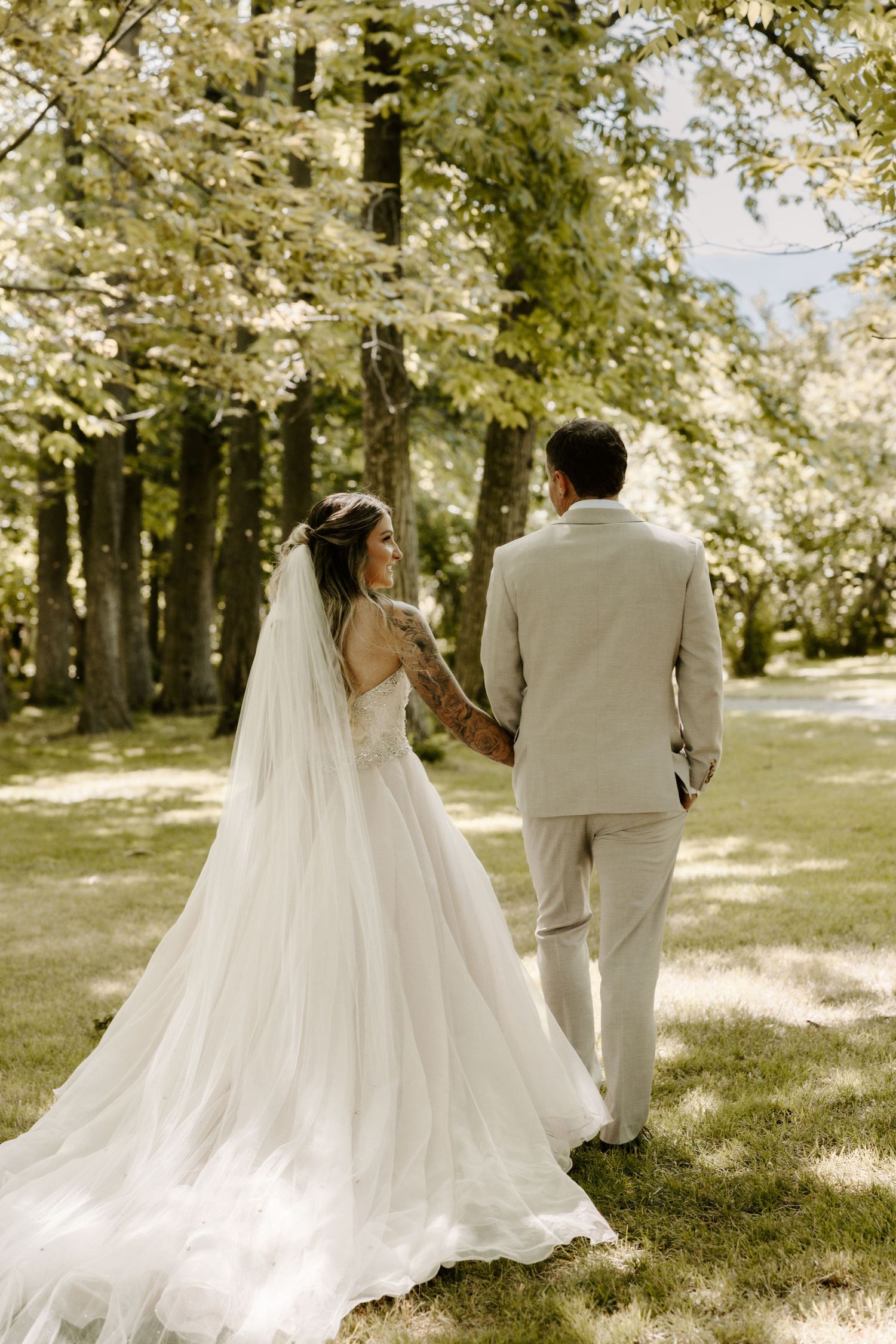 A bride in flowing white gown and groom in light suit walk together through a sunny wooded garden setting.