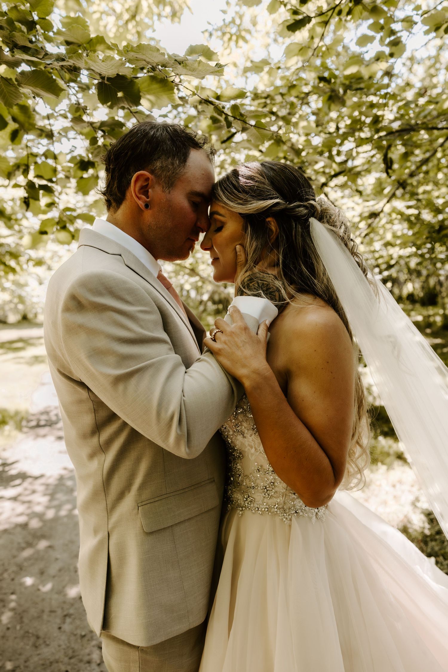 A romantic wedding couple embraces in a sunlit garden setting with the bride wearing a flowing white gown and veil.