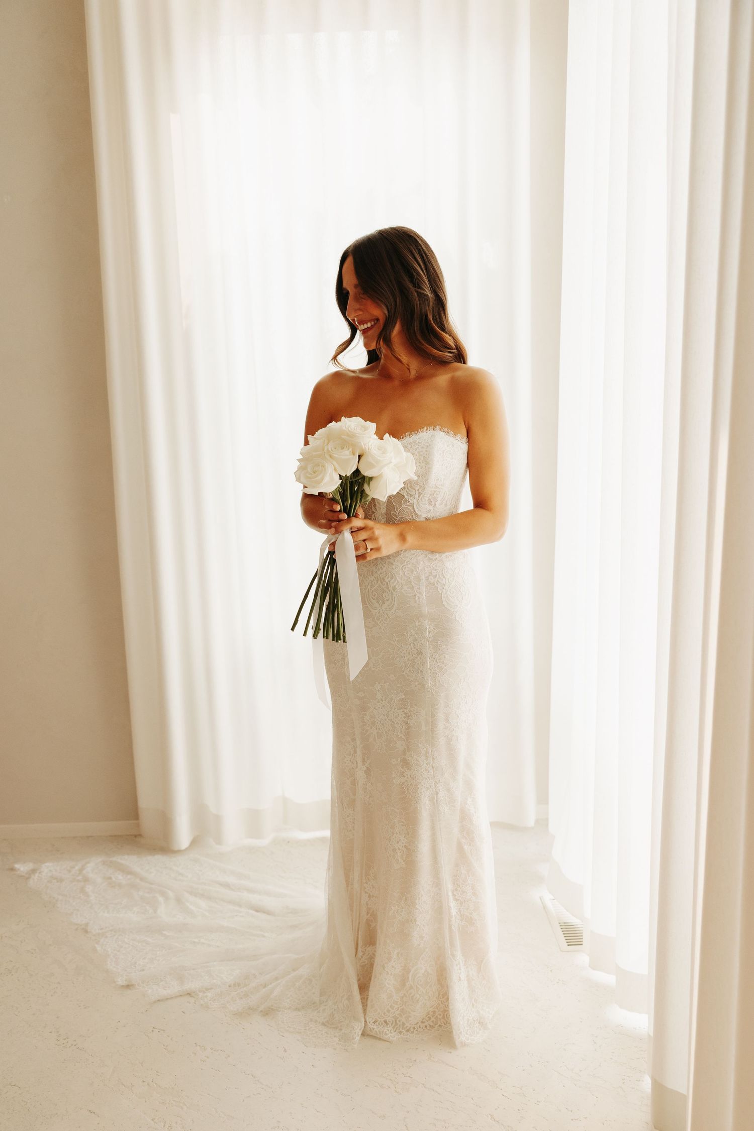 A bride in a white lace wedding gown stands elegantly against sheer white curtains in a bright, sunlit room.