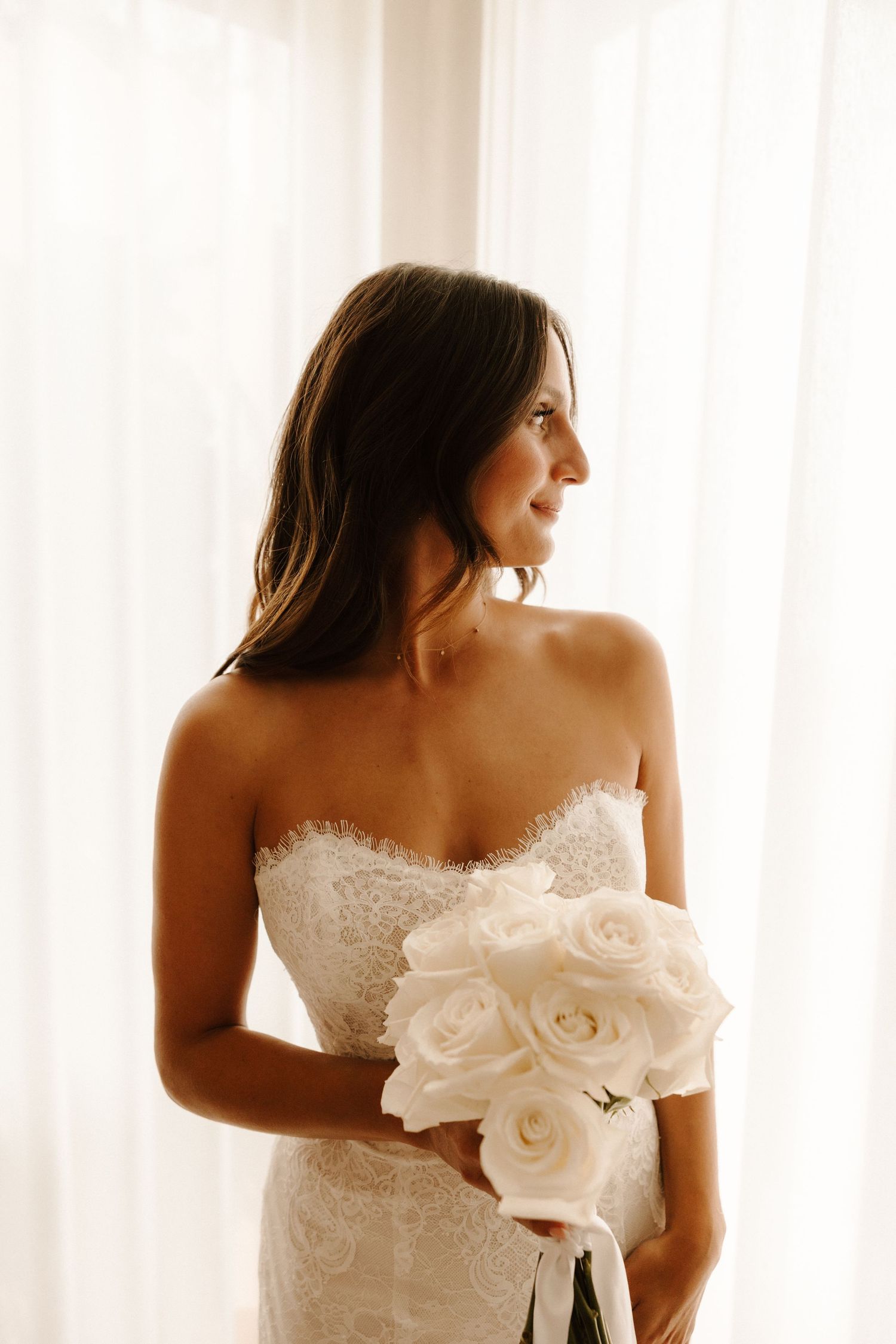A bride in a white strapless wedding gown with delicate lace details poses in soft window light.