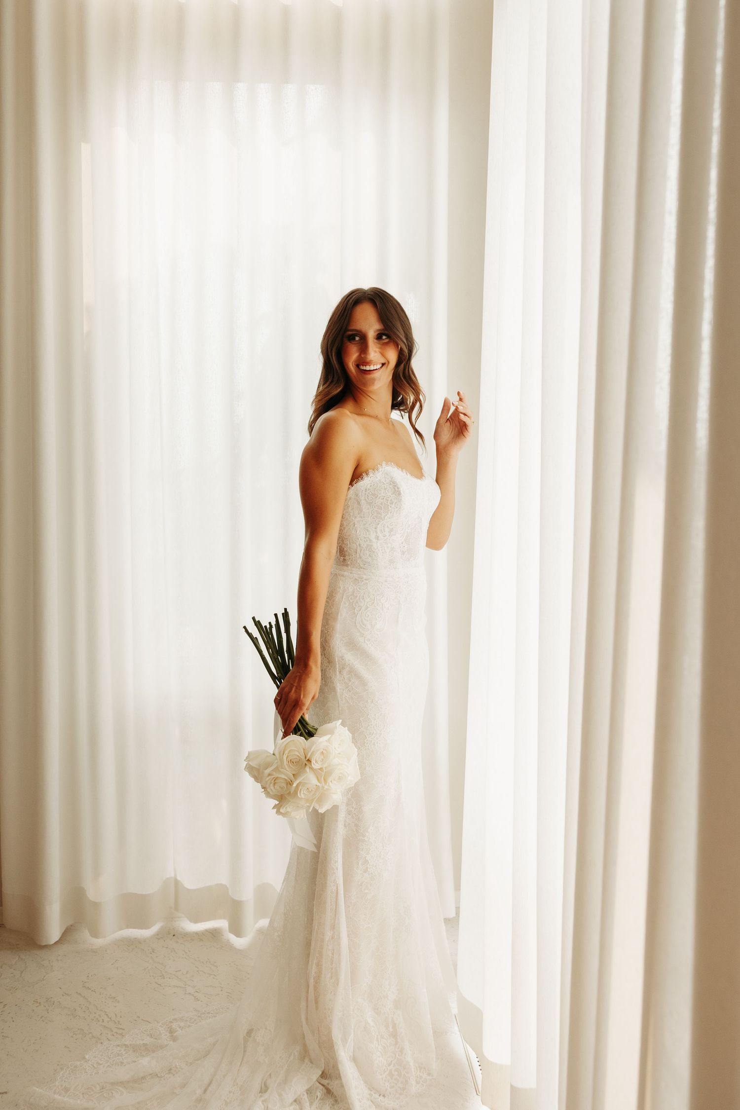 A bride in an elegant white wedding dress poses gracefully against white curtains in a sunlit room.