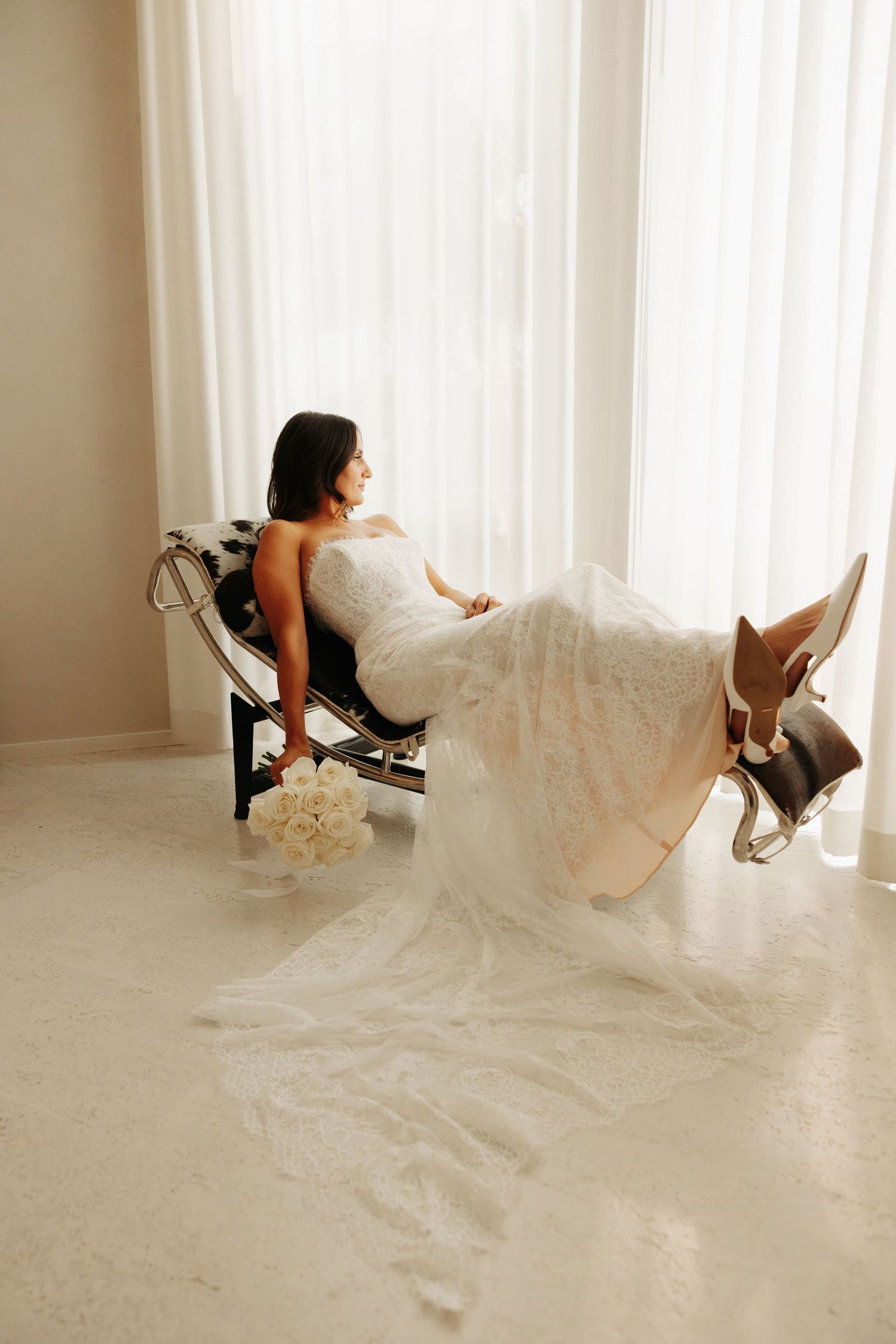 A person in a flowing white wedding dress relaxes in a modern chair with cowboy boots propped up near bright windows.