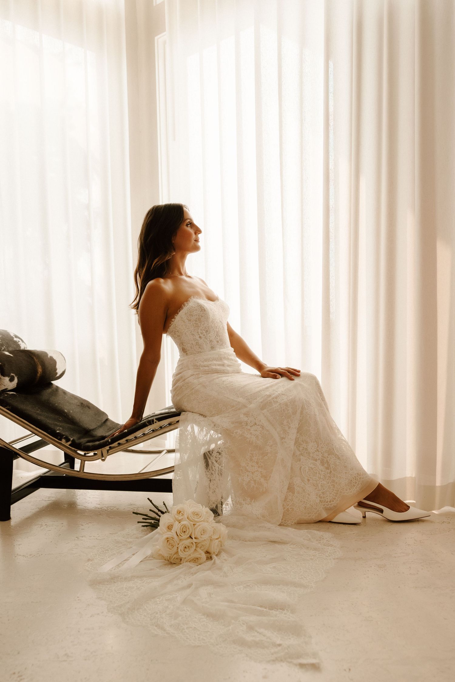 A bride in a white wedding dress reclines on a chaise lounge chair against a light curtain backdrop.