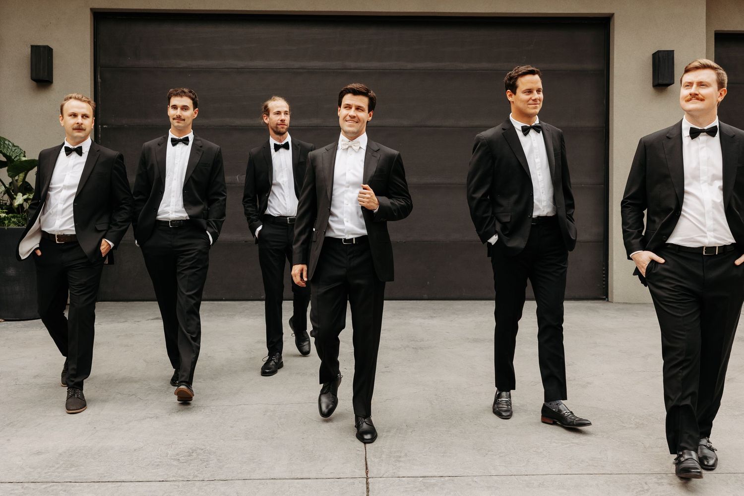 A group of people in formal black tuxedos walk together in a line against a neutral background.