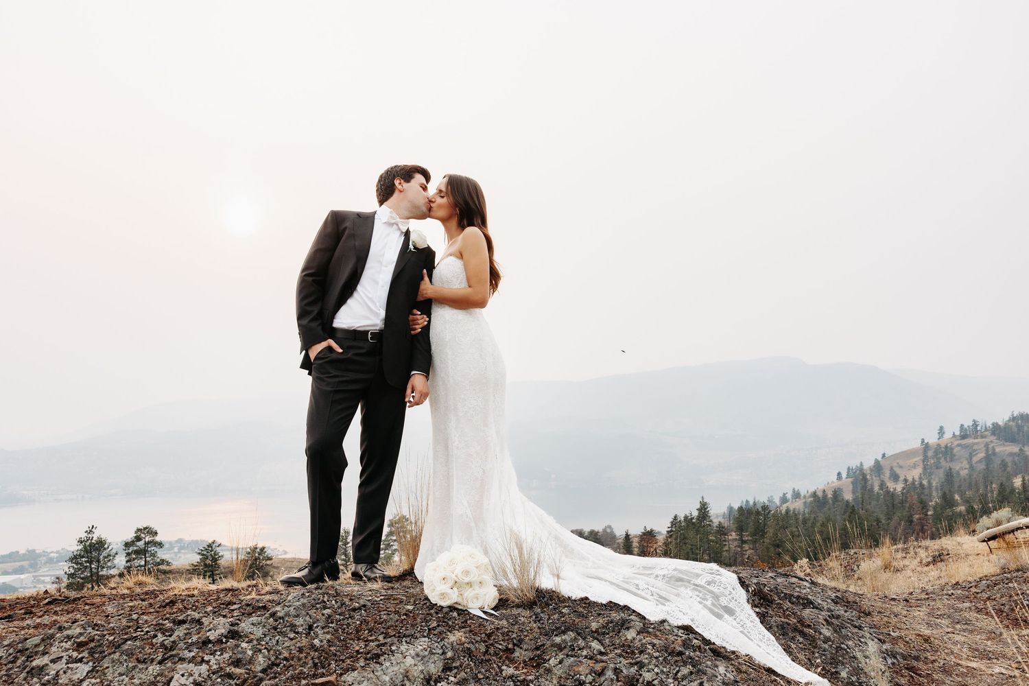 A couple in wedding attire embraces on a mountain summit with scenic mountain ranges in the background at sunset.