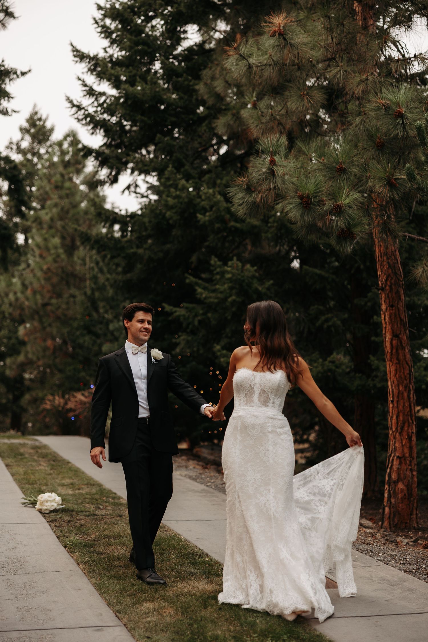 A couple in formal wedding attire walks along a tree-lined path during a romantic outdoor photo session.