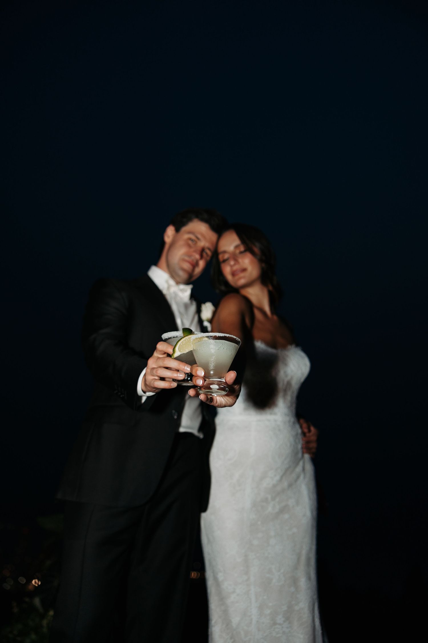 A bride in white lace gown and groom in black tuxedo share a romantic nighttime moment with champagne glasses.