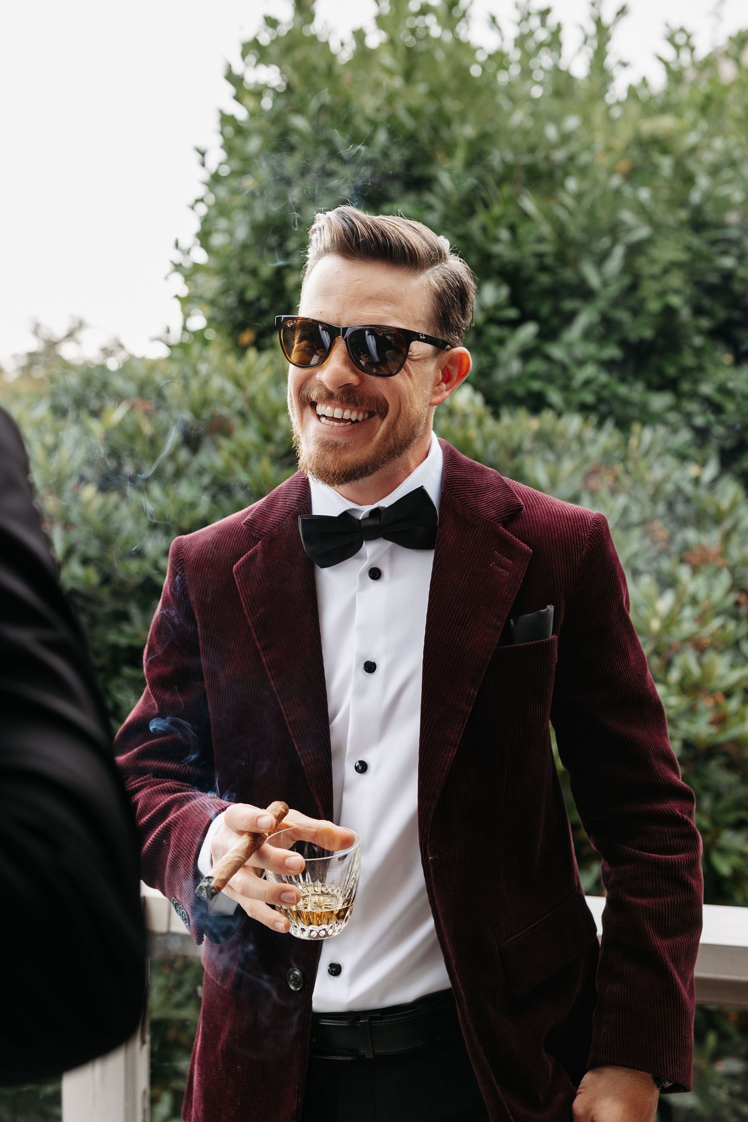 A person in a burgundy velvet tuxedo jacket and black bow tie smiles while holding a drink outdoors.