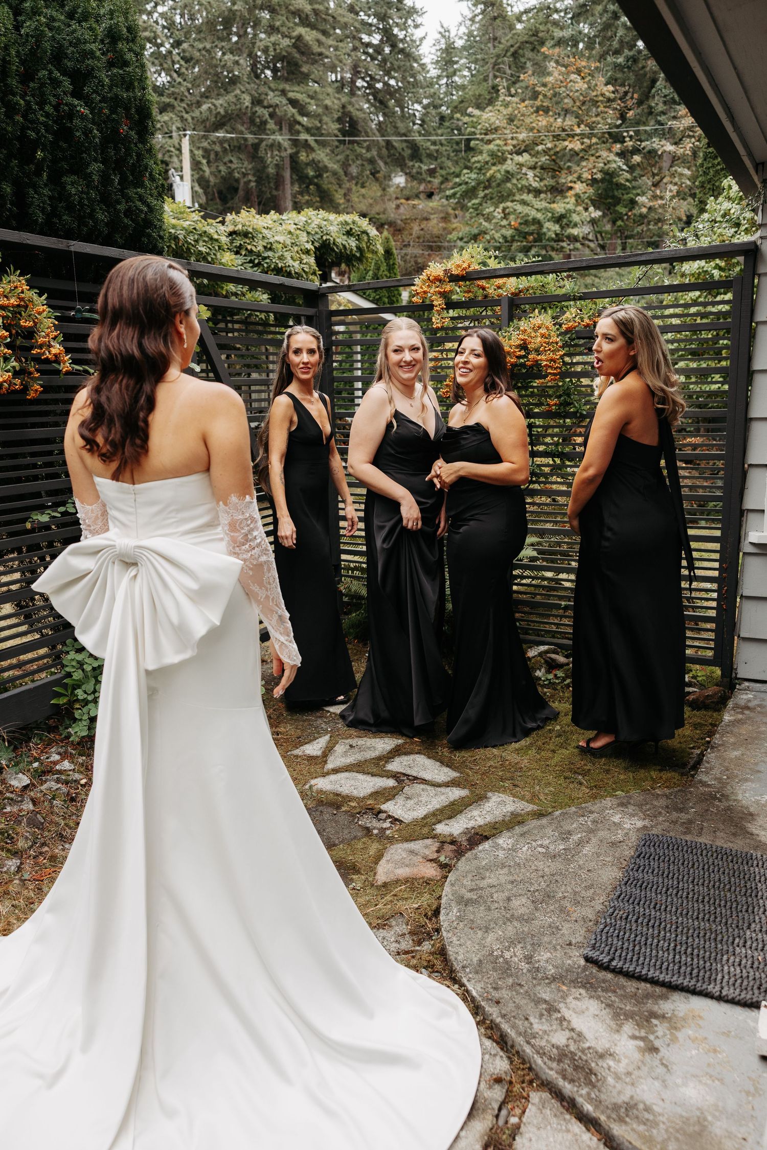 A bride in a white gown with a dramatic bow detail stands with bridesmaids wearing elegant black dresses in an outdoor garden setting.