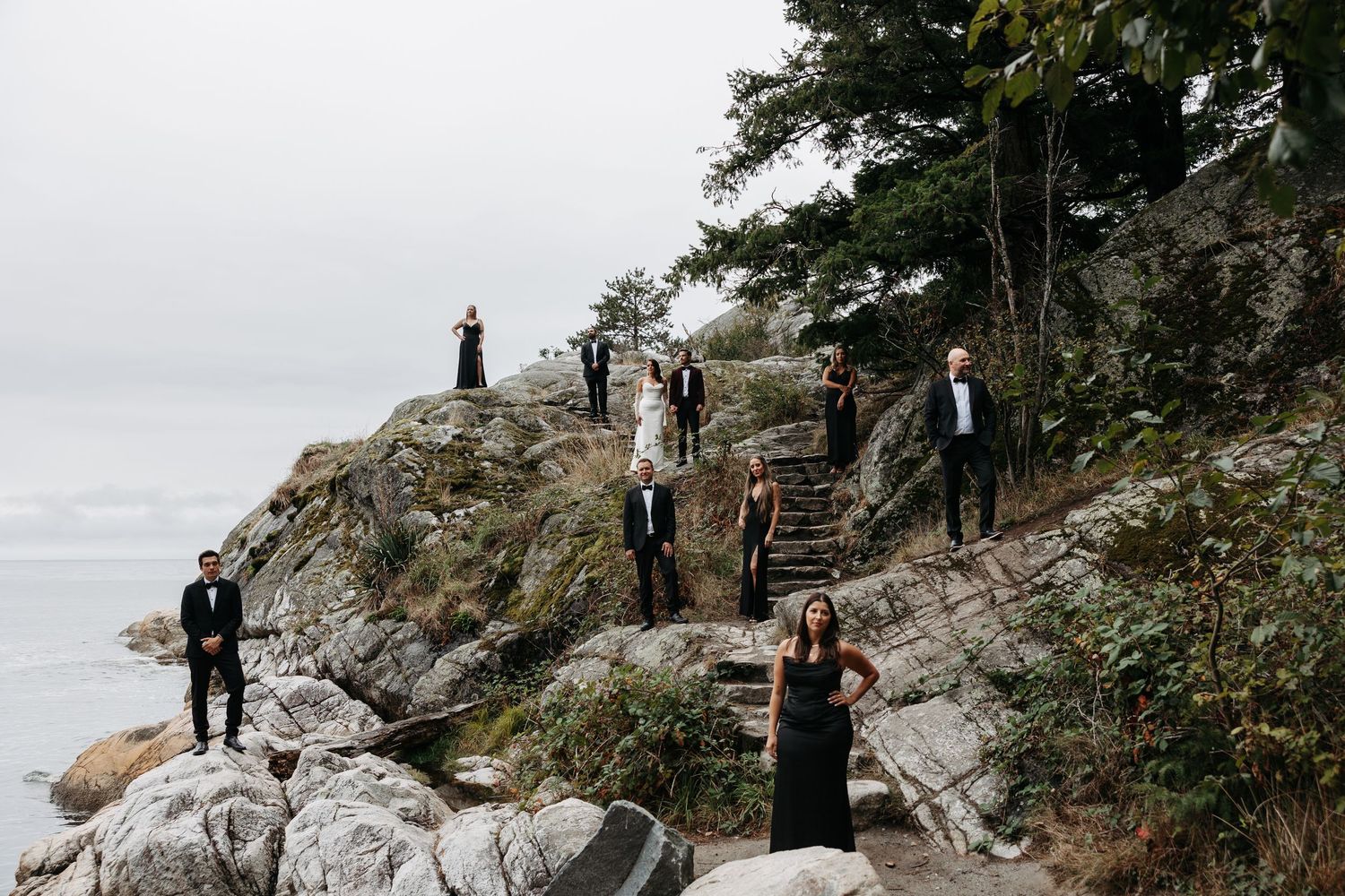 A group in formal attire stands on rocky coastal cliffs with wooden stairs and evergreen trees in the background.