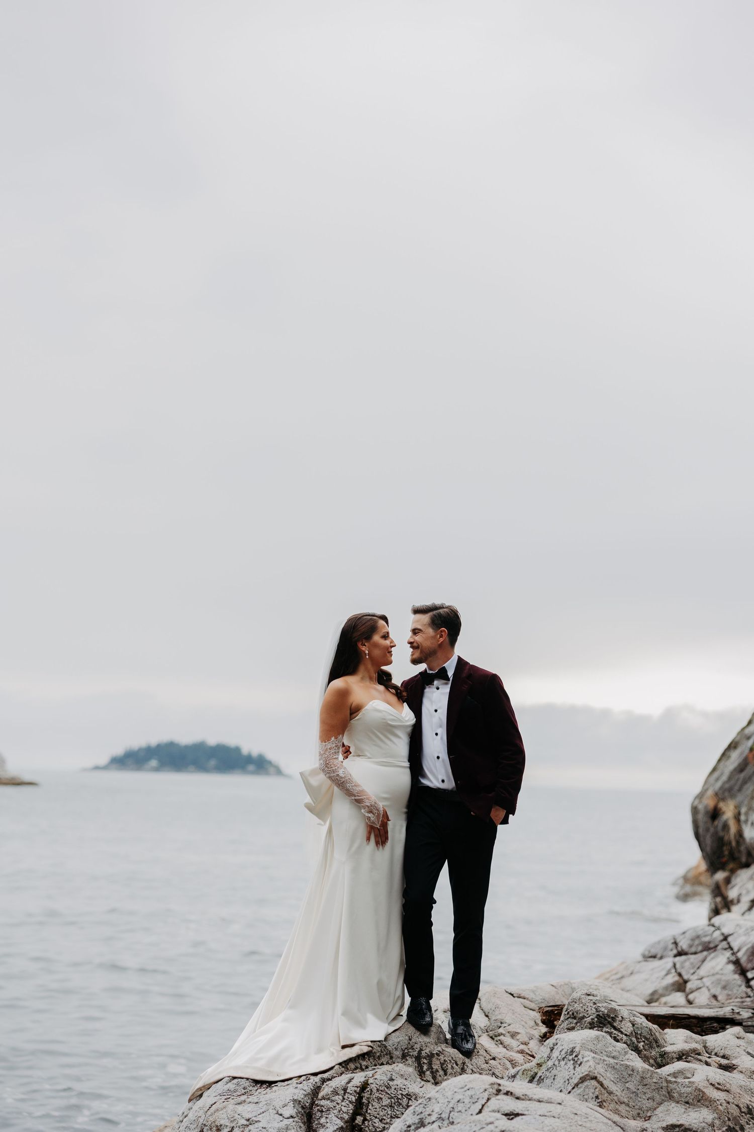 A romantic wedding couple embraces on rocky coastal cliffs overlooking a misty ocean view.