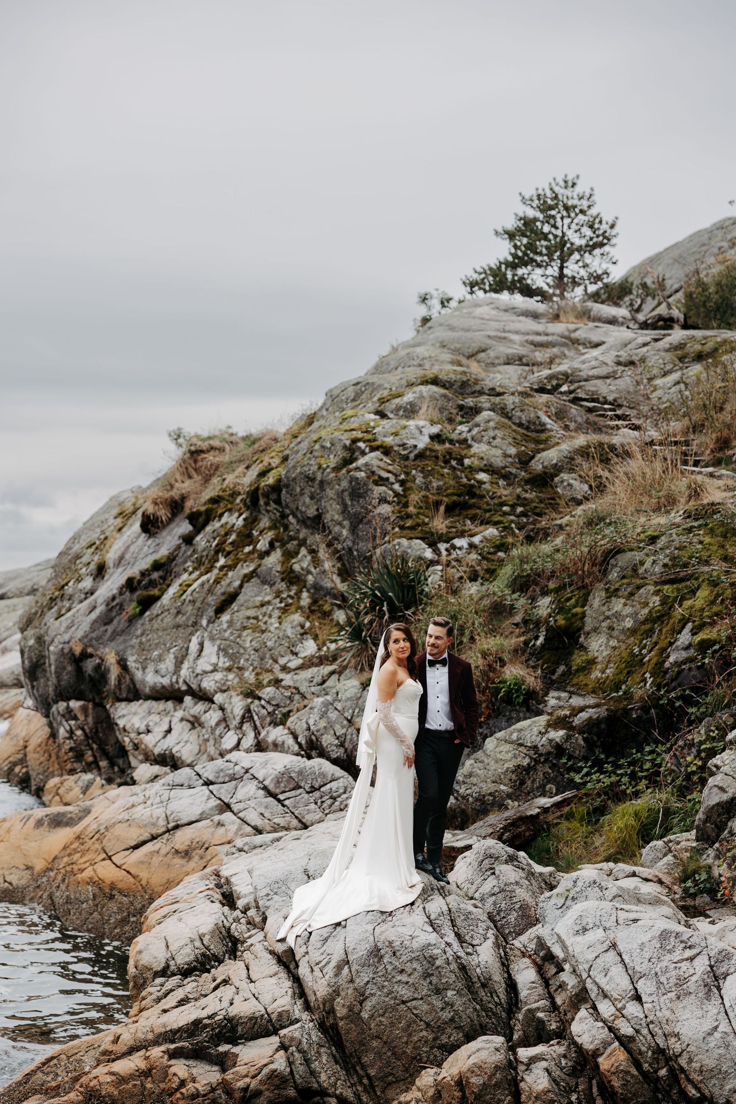 Newlywed couple poses on dramatic rocky coastal cliffs overlooking the Pacific Ocean on a cloudy day.