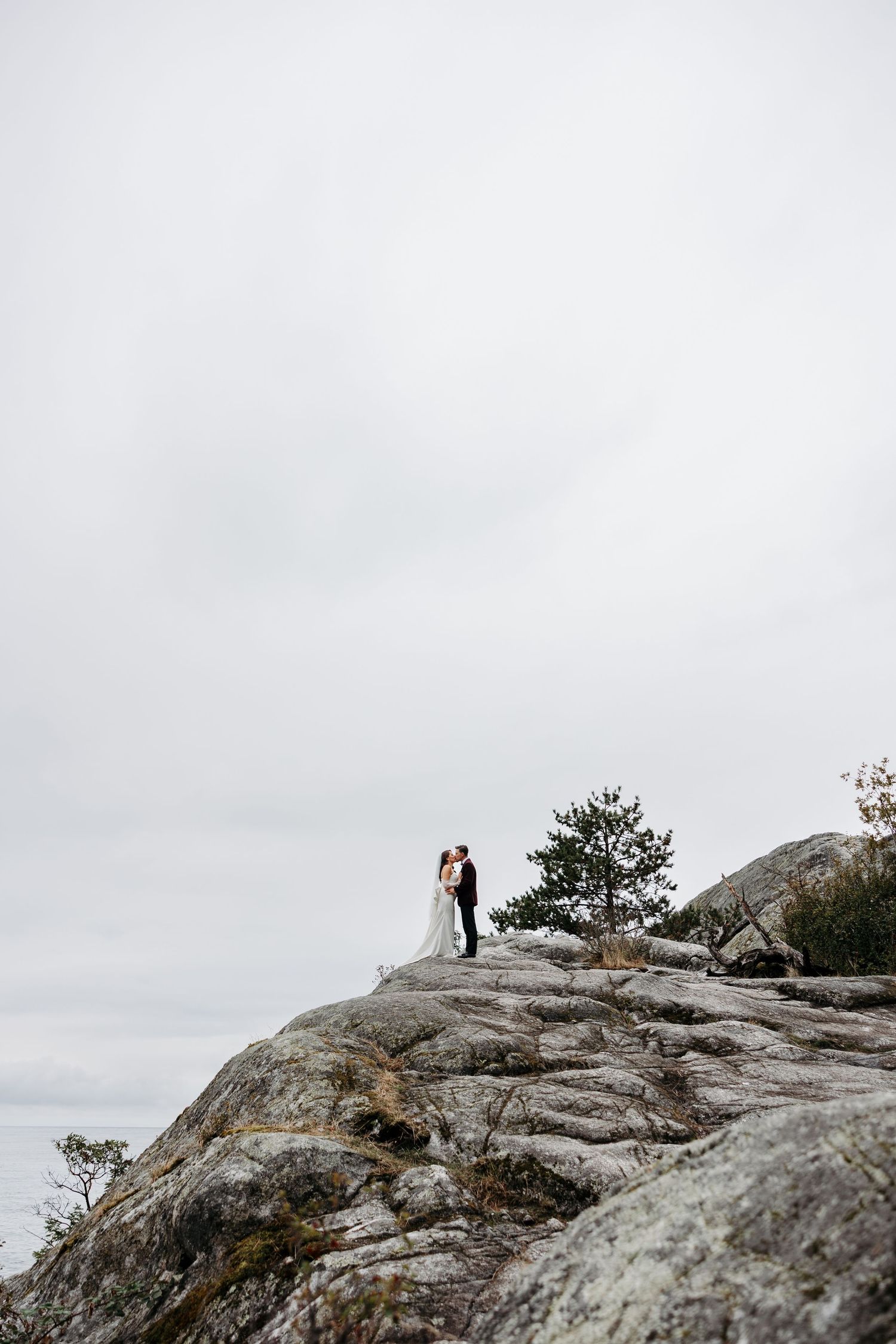 Two figures stand atop a rocky mountain peak against a cloudy sky during what appears to be a wedding ceremony.