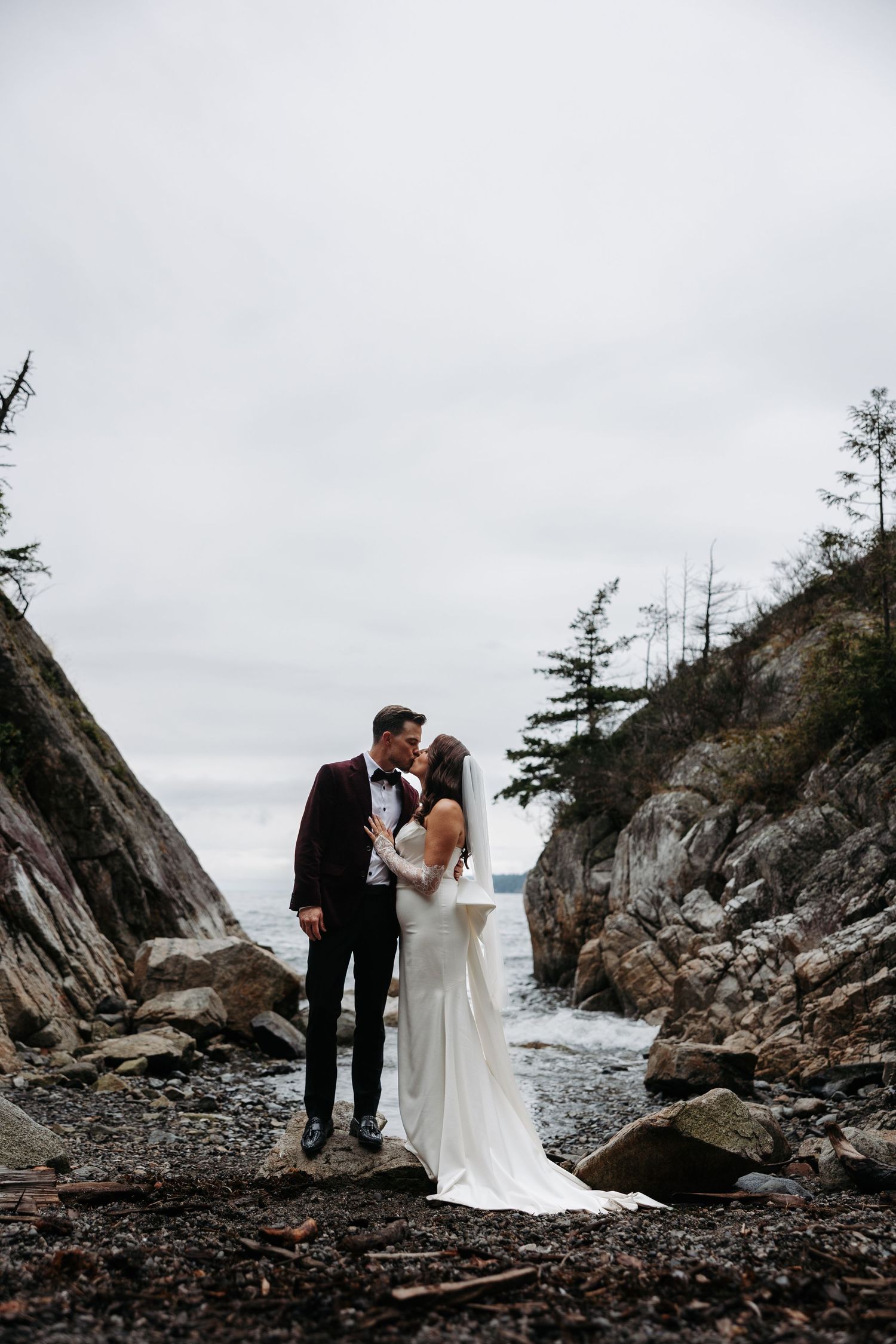 A couple shares an intimate moment on a rocky Pacific Northwest beach with dramatic cliffs and driftwood in the background.