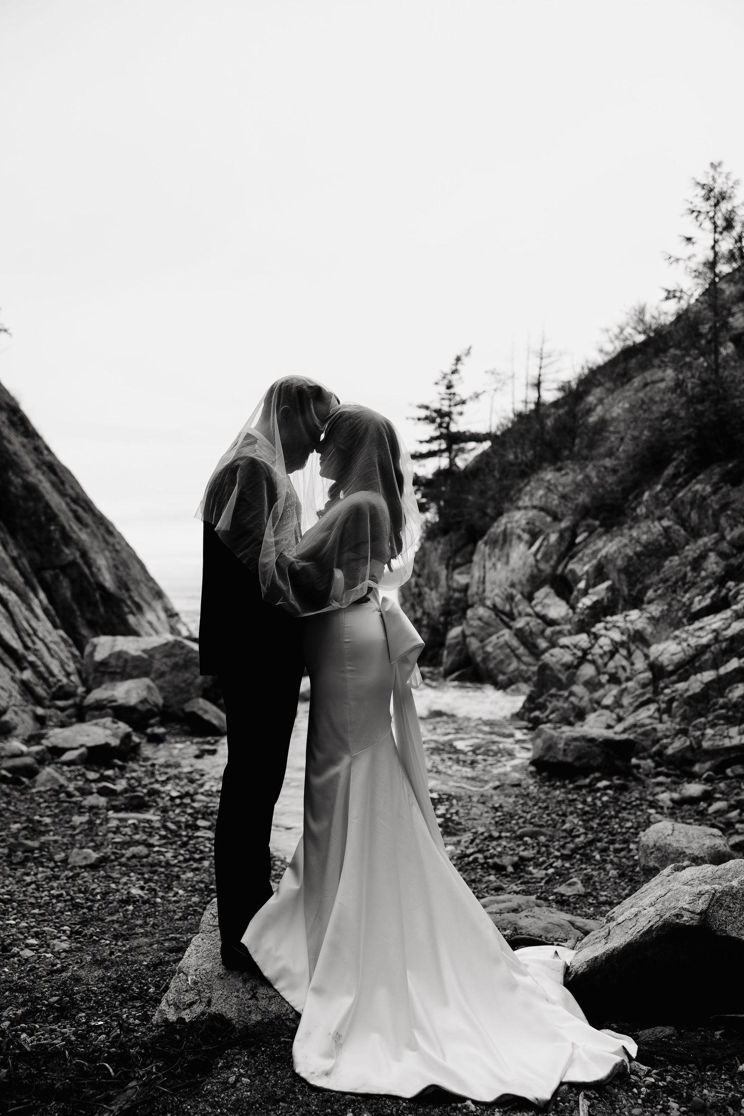 A romantic black and white wedding photo of a couple embracing on a rocky beach surrounded by cliffs.