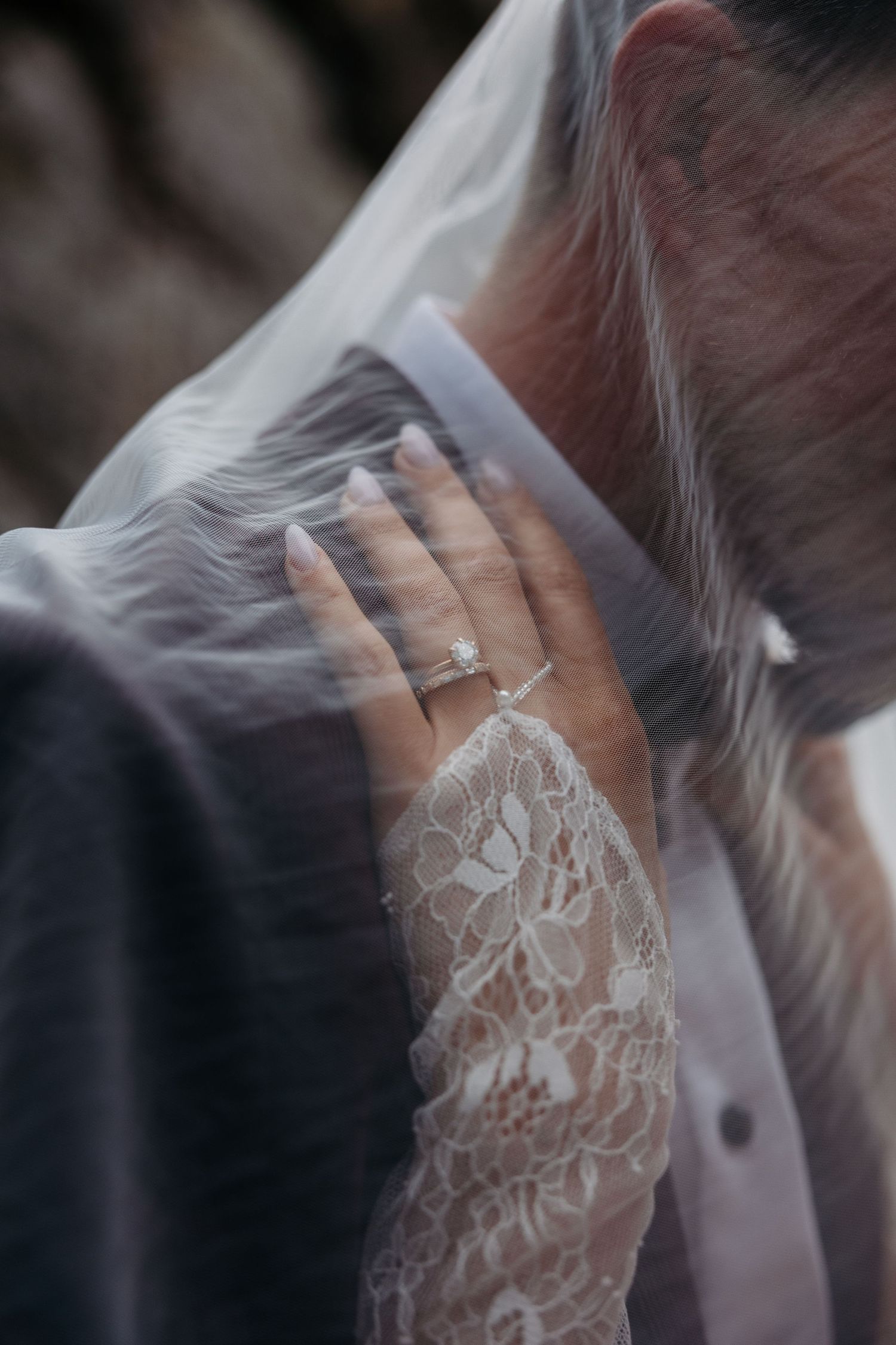 Close-up of delicate white lace sleeve and wedding ring through sheer bridal veil in soft lighting.