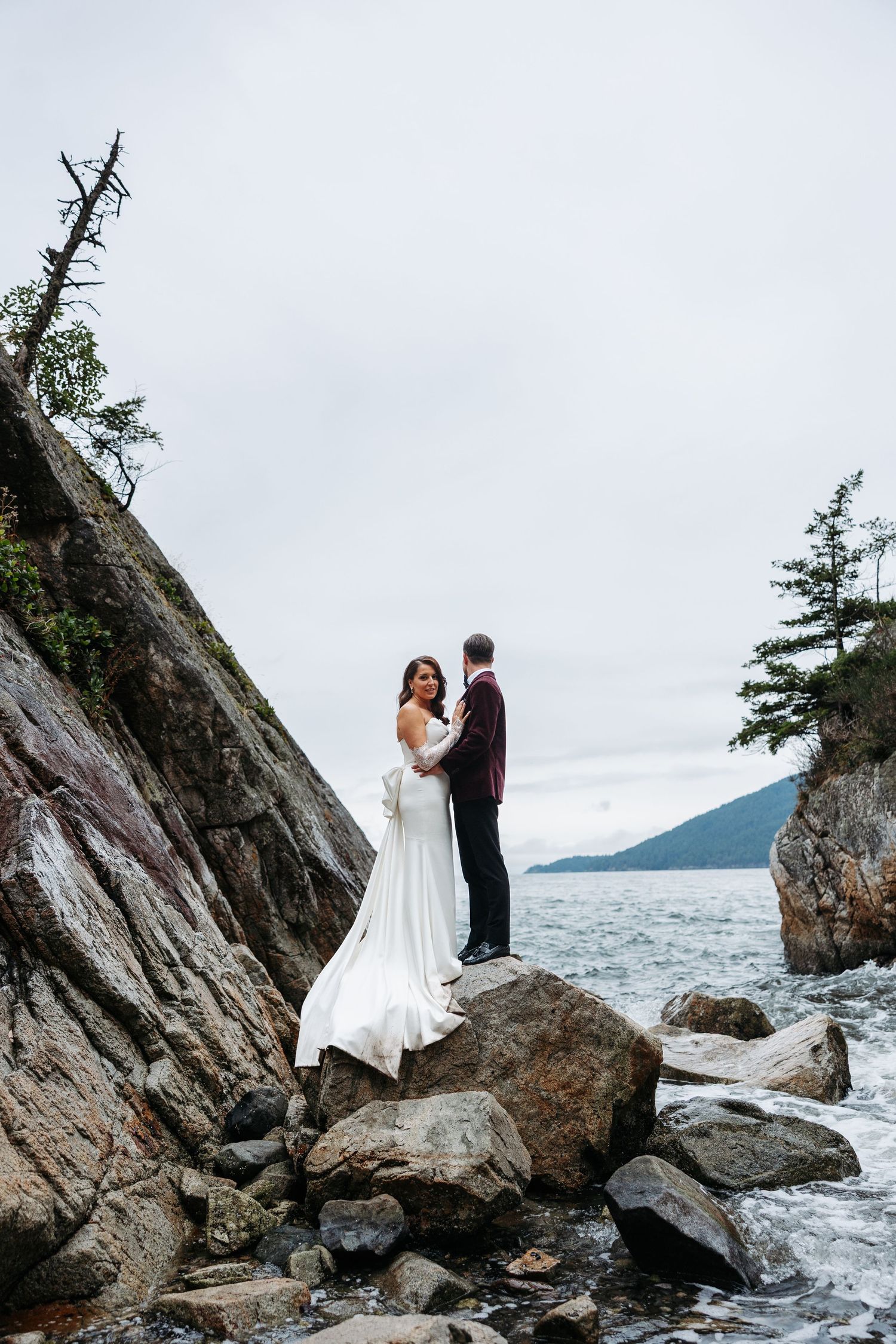 A couple shares an intimate moment on rocky coastal cliffs overlooking the ocean and mountains on their wedding day.