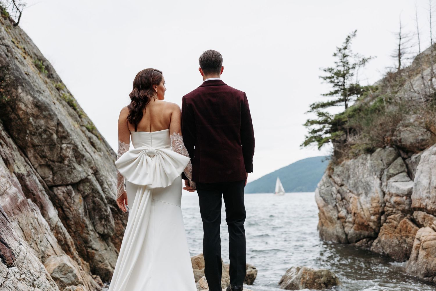 A couple in wedding attire walks along a rocky coastal cliff with ocean views in the background.