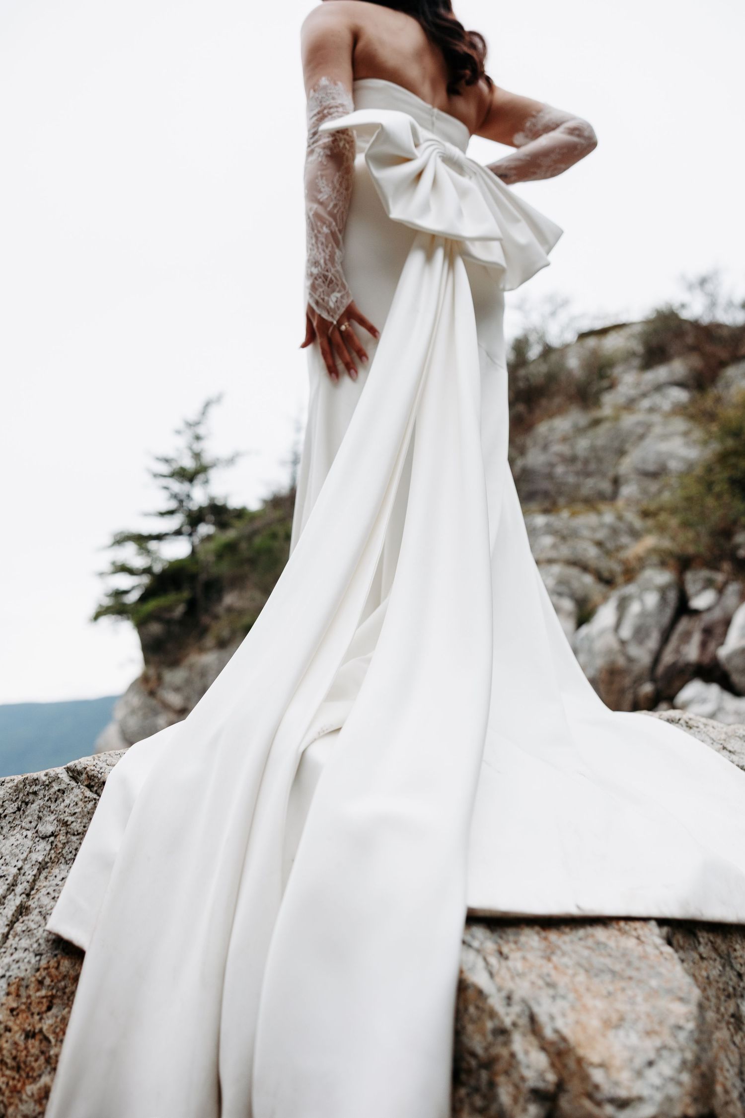 A flowing white wedding dress with lace details and dramatic bow billows against a rocky coastal backdrop.