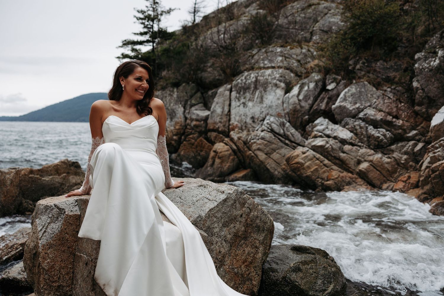 A bride in a flowing white dress poses gracefully on rugged coastal rocks with crashing waves in the background.