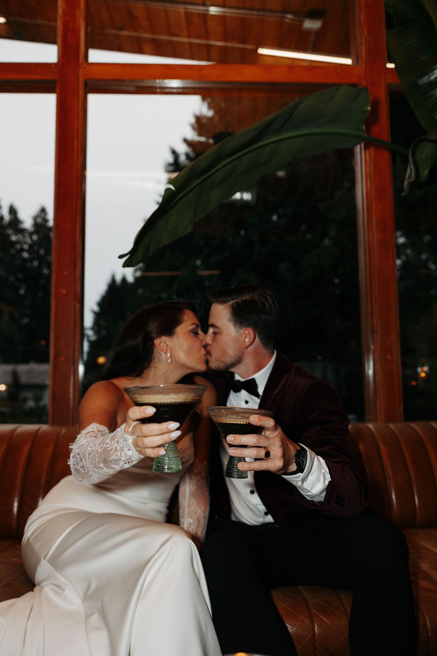 A couple in wedding attire shares a romantic toast while seated in a rustic venue with wooden paneling.