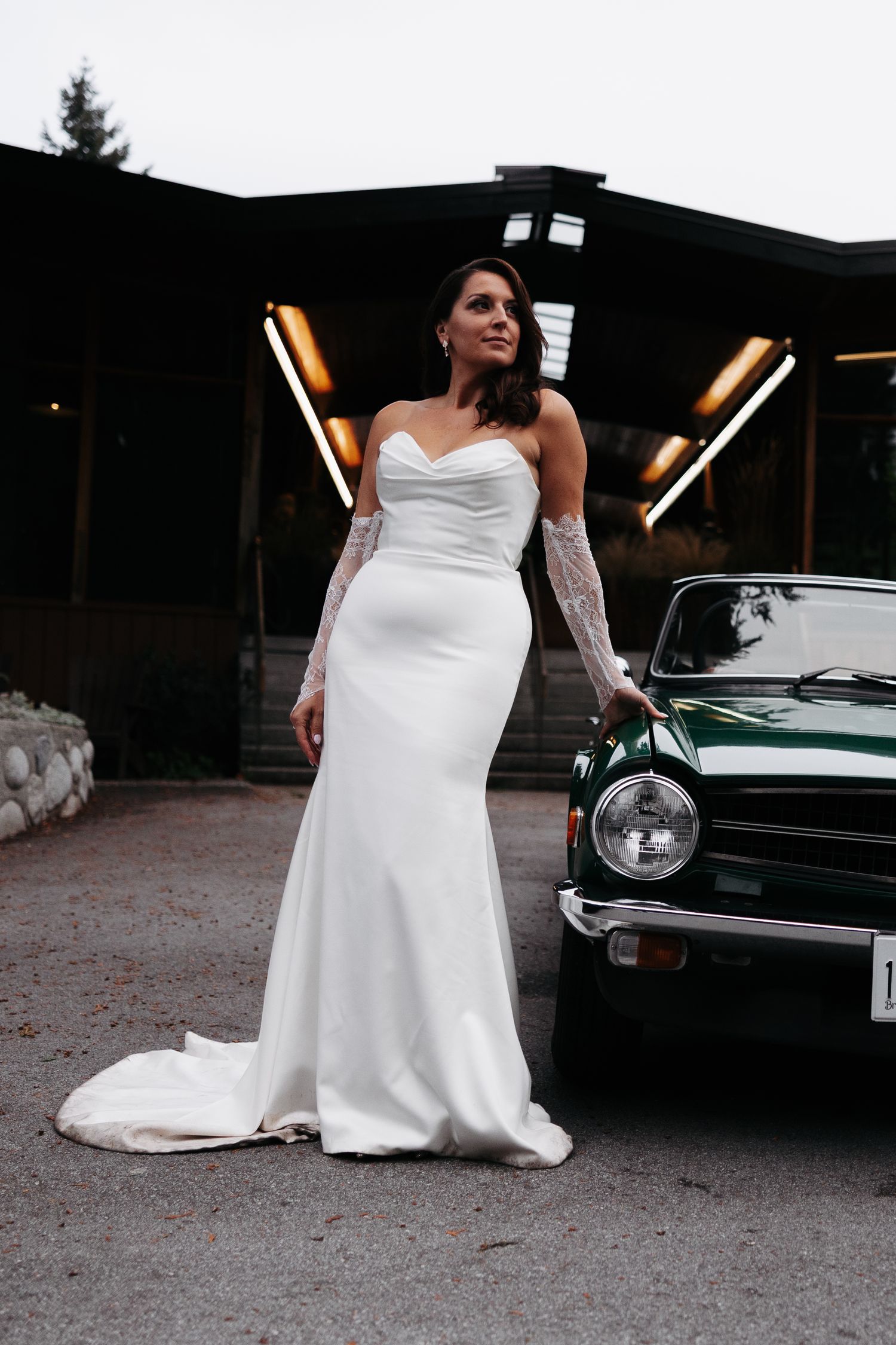 A bride in a white wedding gown poses elegantly next to a vintage car during a photoshoot.
