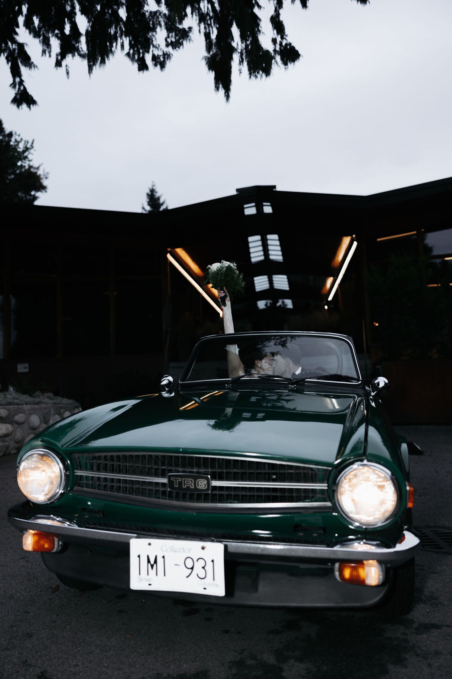 Vintage green sports car with headlights on parked under trees at dusk.
