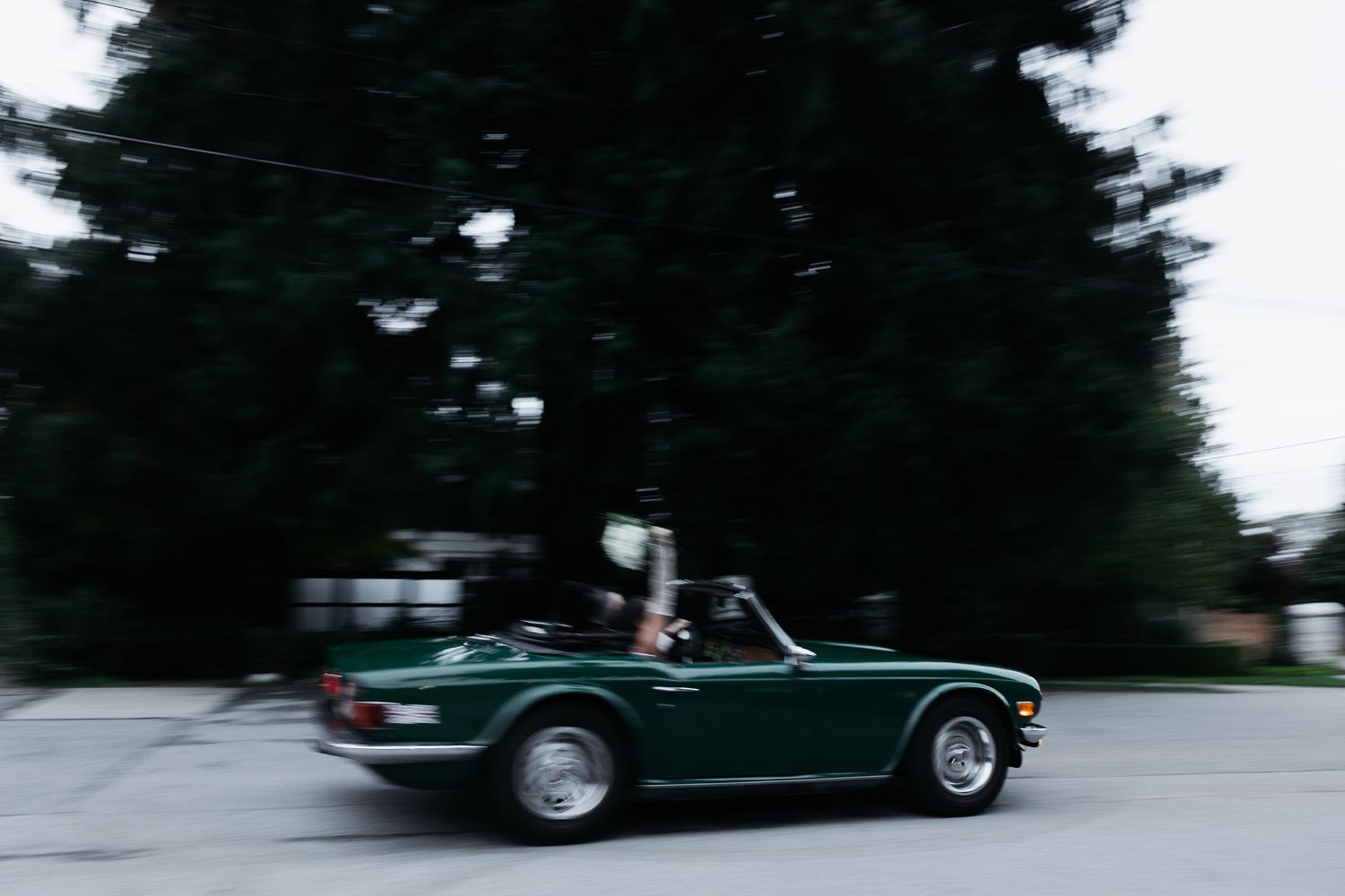 Blurred motion shot of dark green classic car driving on tree-lined road.