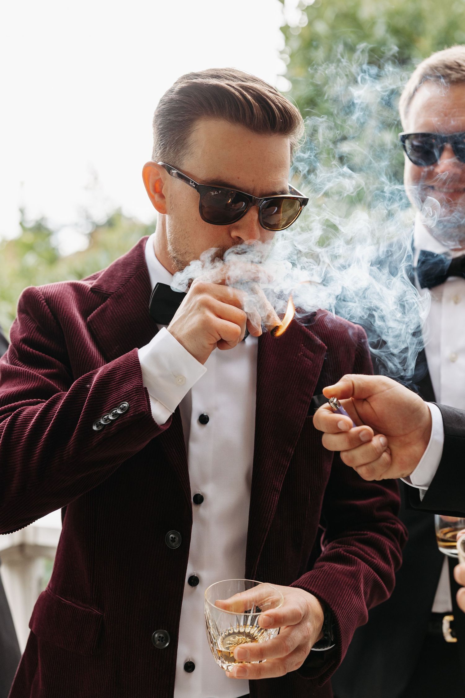 A person in a burgundy suit jacket and white shirt lights a cigar while holding a glass of whiskey.