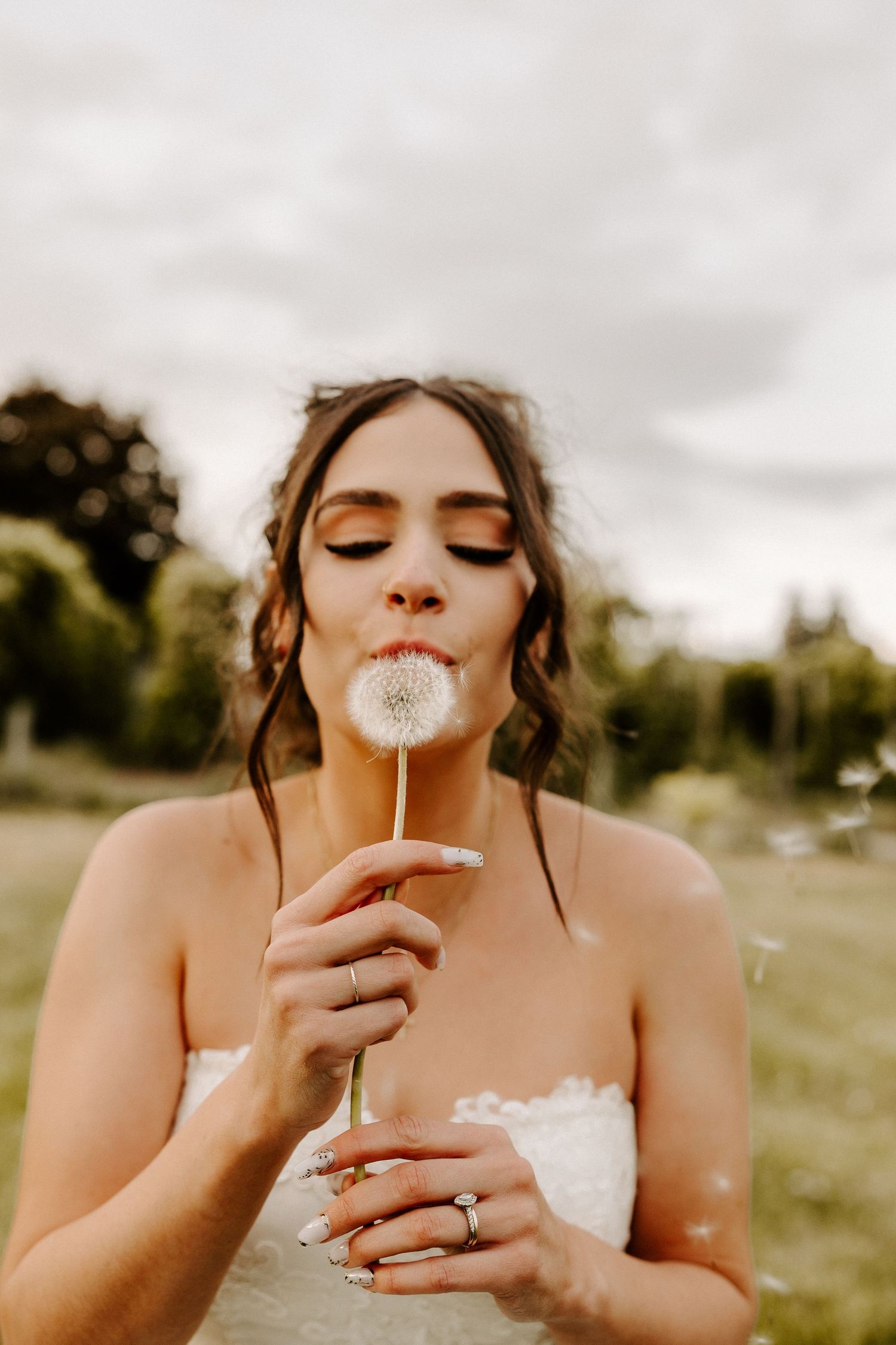 A bride in a white dress blows on a dandelion in a meadow during a golden hour outdoor photoshoot.