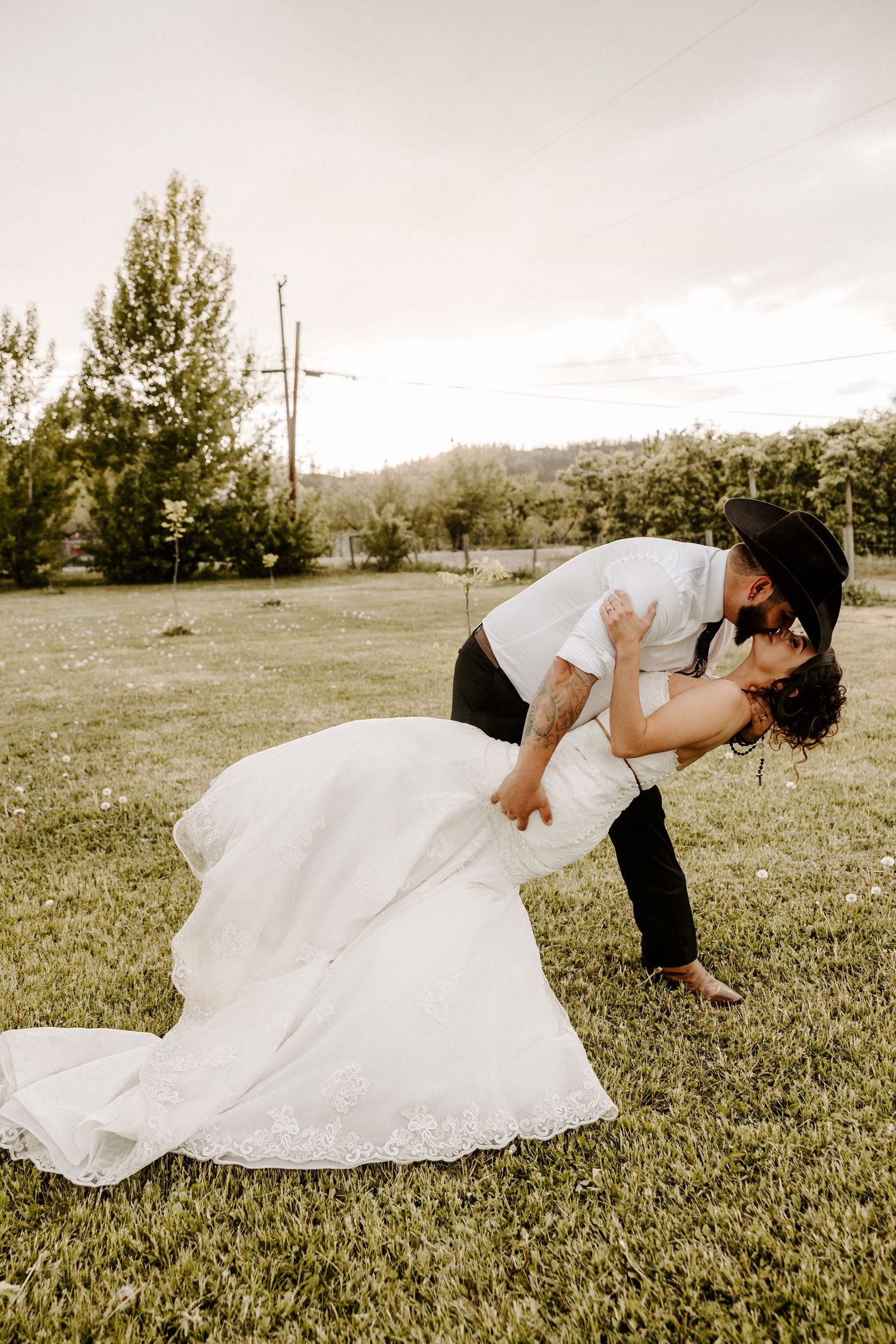 A romantic wedding couple embraces in a dip pose on a grassy field with scenic mountains and trees in the background.