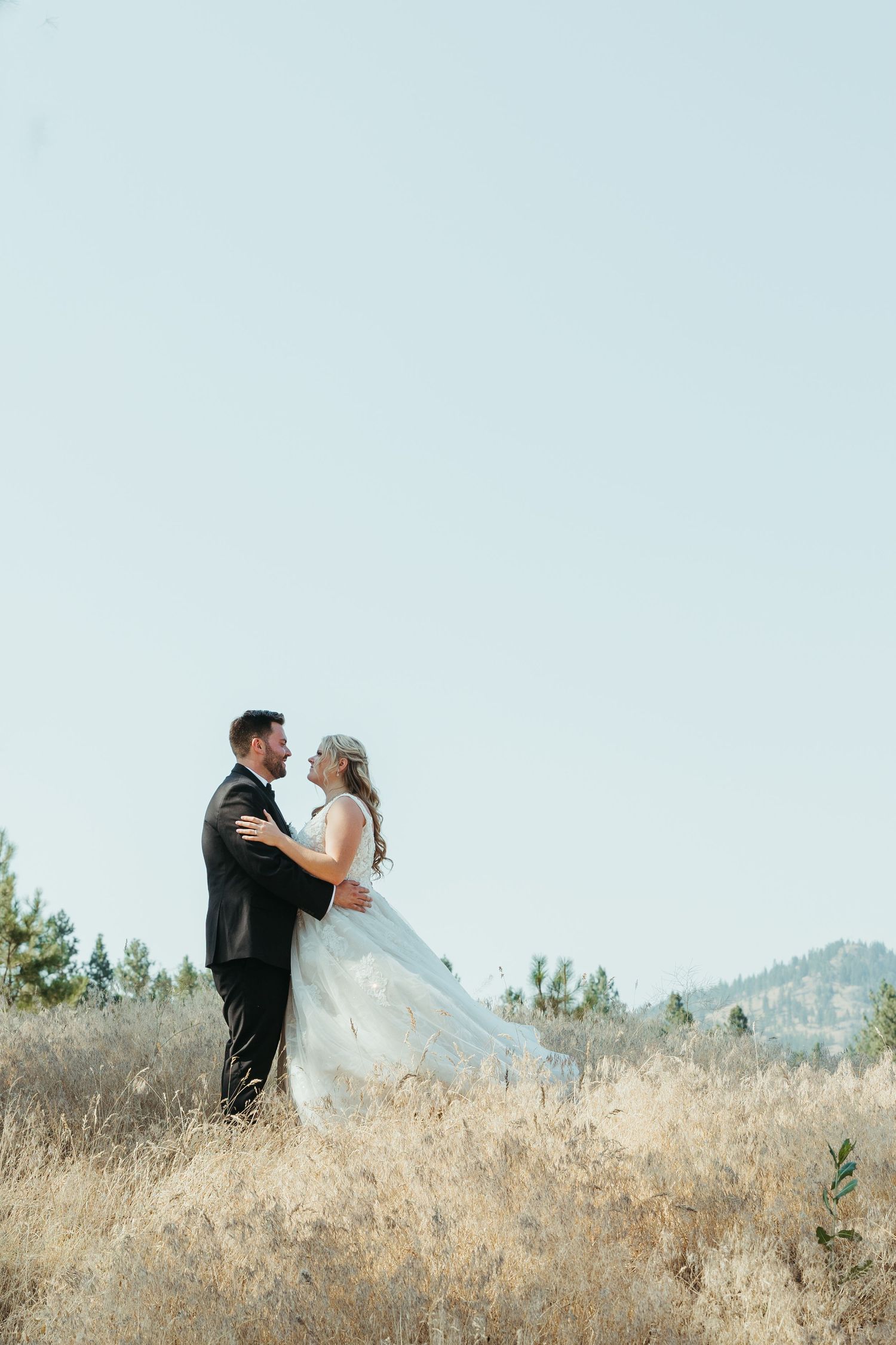 Bride and groom embrace on hilltop with mountains and blue sky in background.