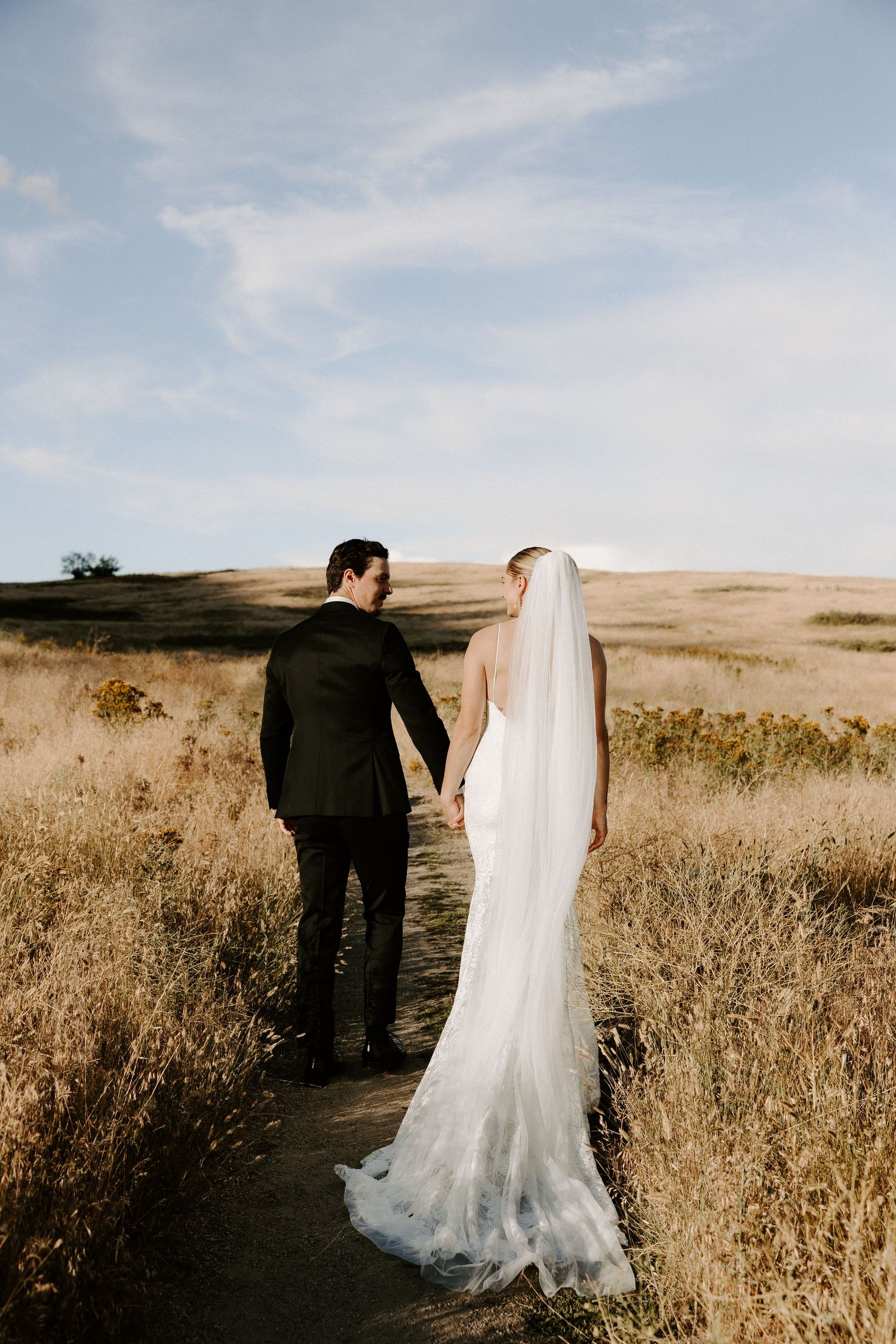 A couple in wedding attire walking down a dirt path through golden hills on their special day.