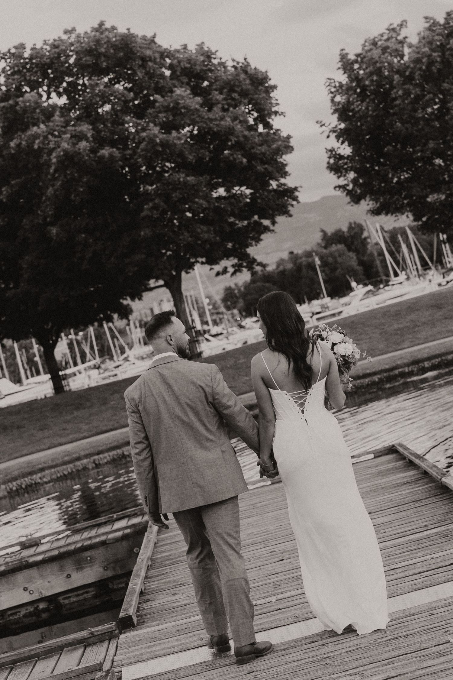 A couple in wedding attire walking together on a wooden dock near sailboats and trees in black and white.