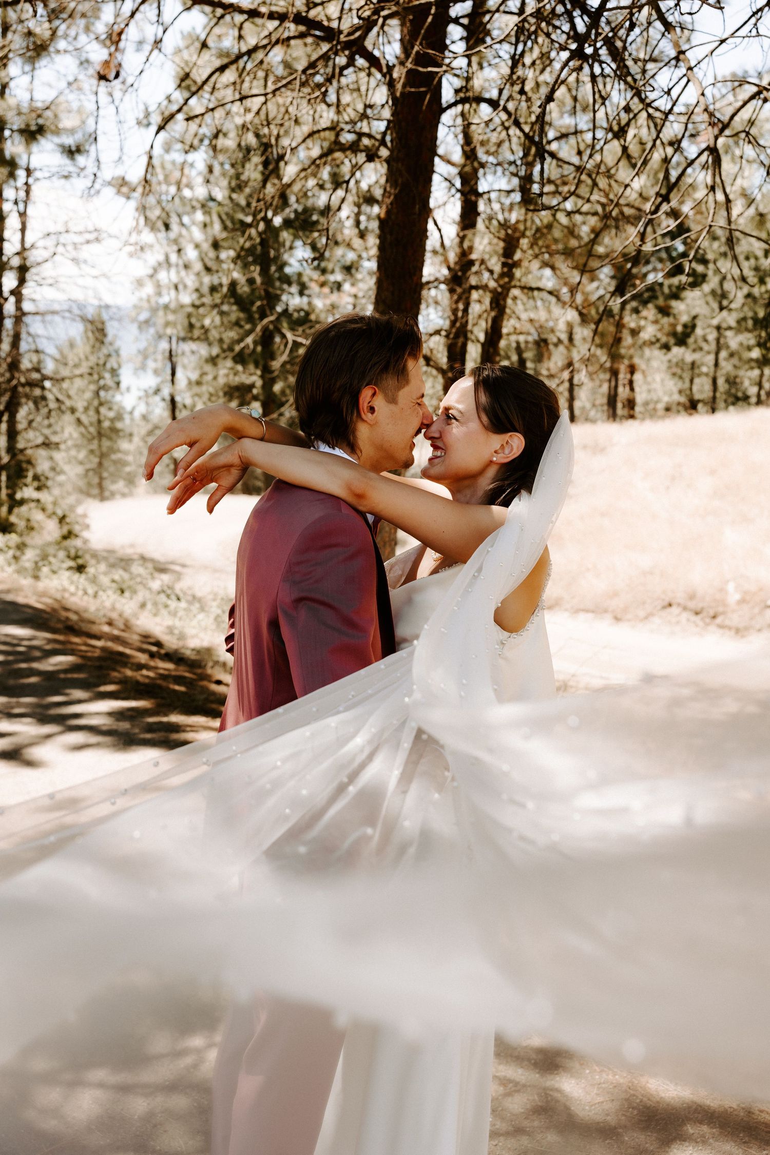 A couple embraces on their wedding day in a sun-dappled forest as the bride's veil flows dramatically in the breeze.